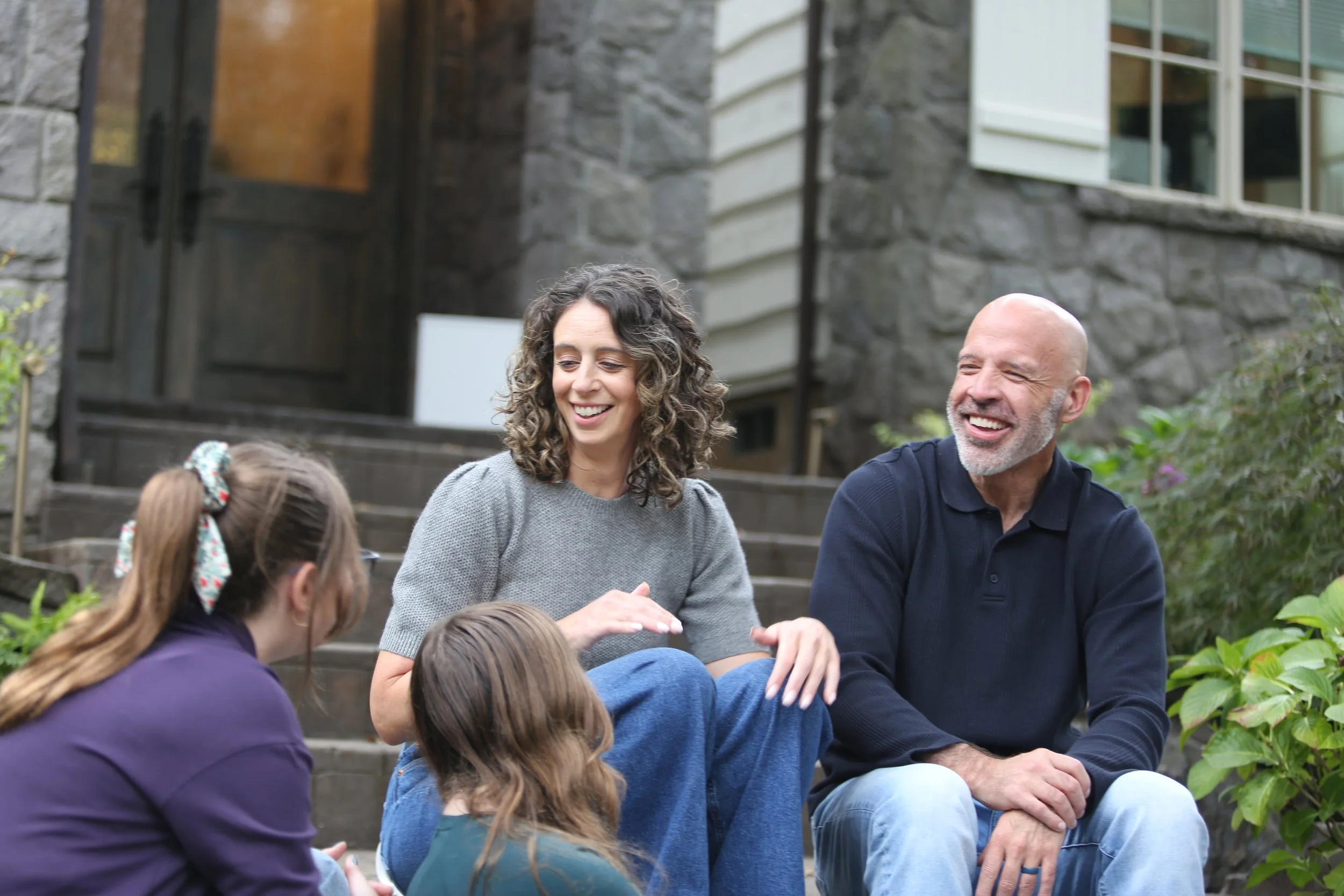 A group of children and adults sitting outdoors on steps with a stone building and greenery in the background, engaged in conversation or activity, smiling and interacting.