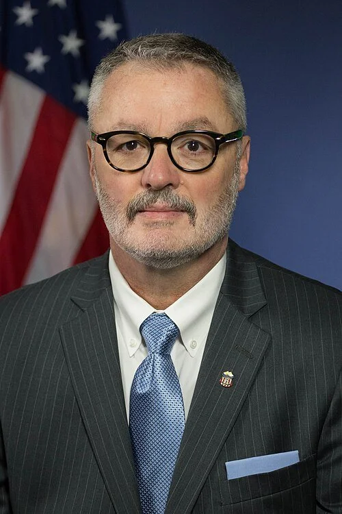 A middle-aged man with short gray hair and a beard, wearing glasses, a dark pinstripe suit, a white shirt, and a blue tie. He stands in front of an American flag backdrop.