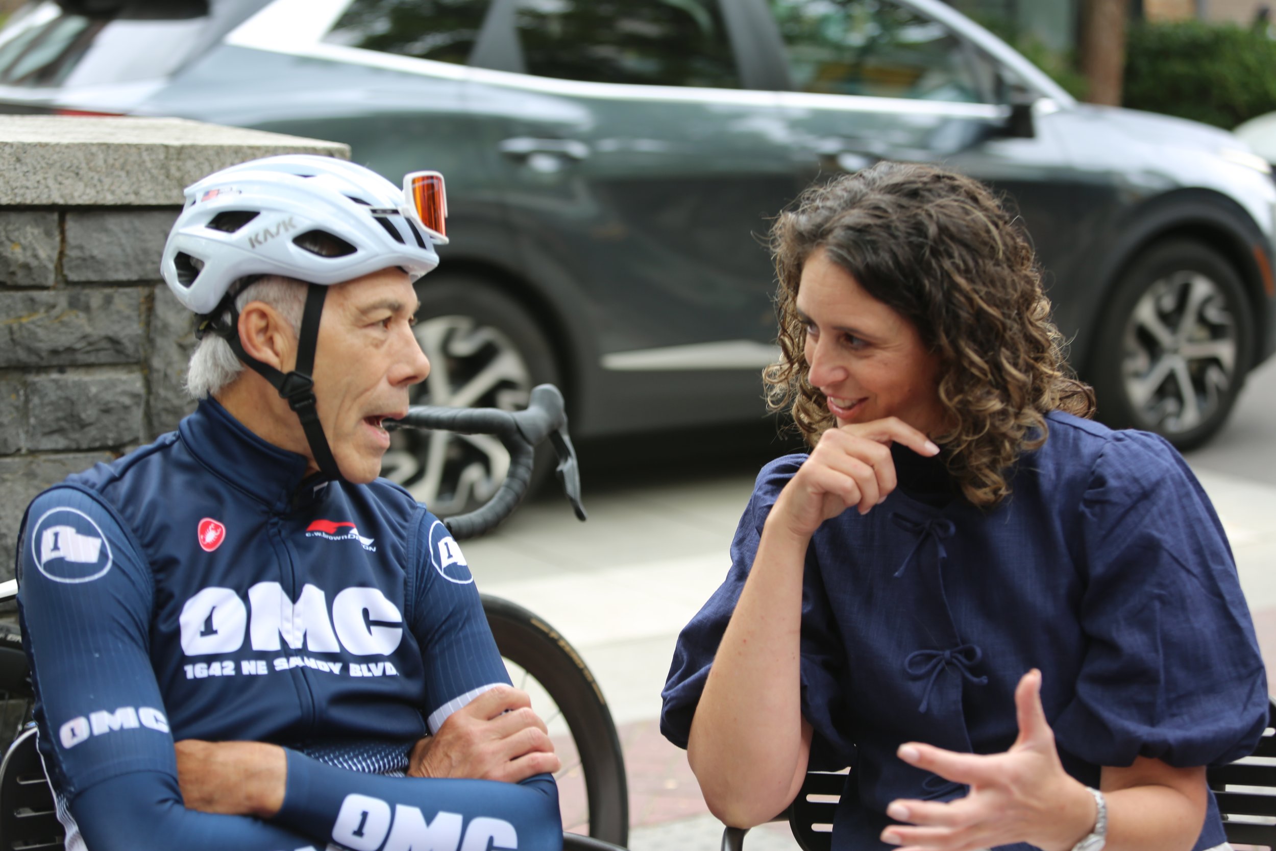 A senior man in cycling attire wearing a helmet and glasses, sitting and talking to a woman with curly hair in a blue jacket, outdoors with cars and trees in the background.
