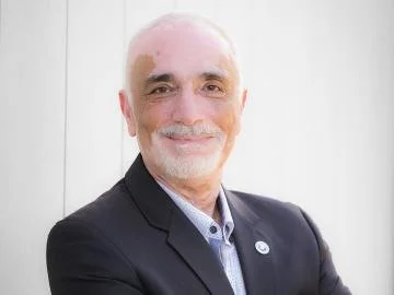 A middle-aged man with a bald head and a white beard wearing a black blazer and a light blue shirt, smiling in front of a light-colored wall.