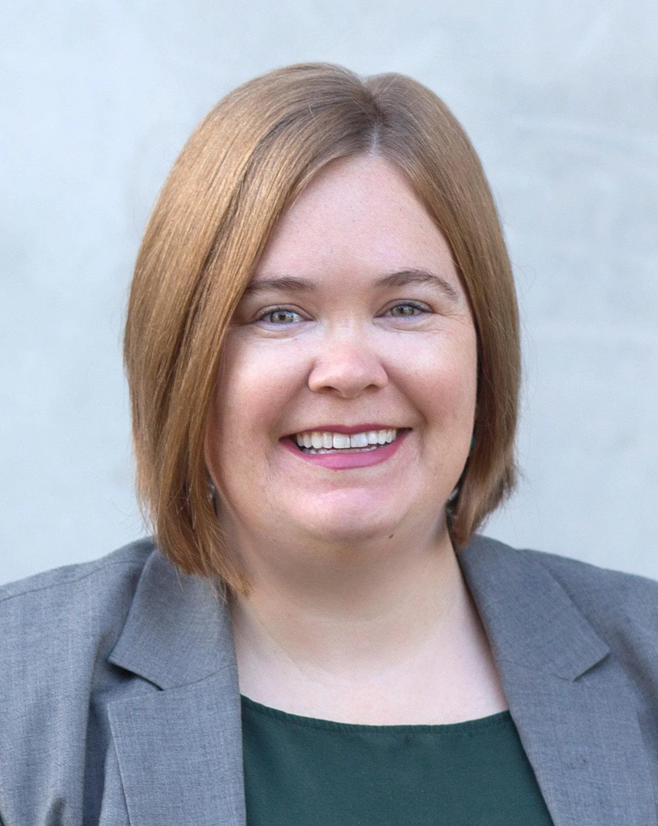 Close-up of a smiling woman with shoulder-length red hair, wearing a grey blazer and a dark top, with a plain light background.