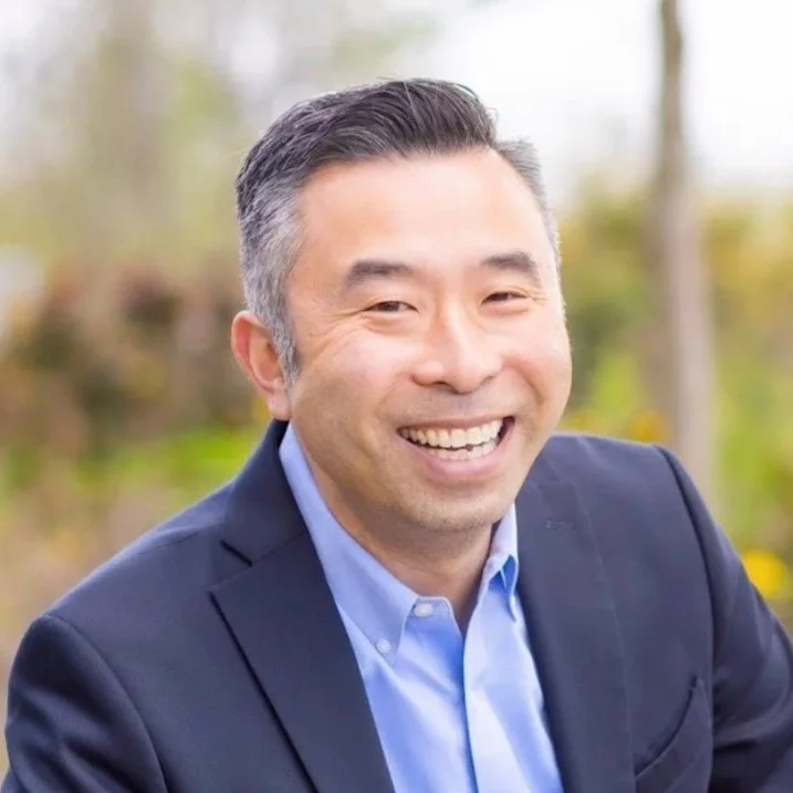 Smiling man in a suit outdoors with blurred trees in the background.