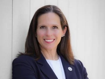 A woman with shoulder-length brown hair, wearing a navy blazer and white top, smiling in front of a light-colored wooden wall.