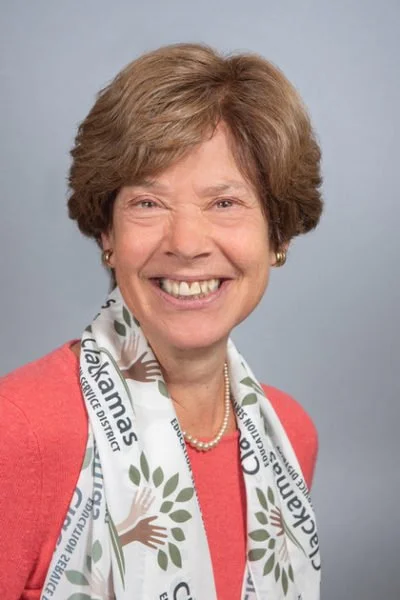 Smiling middle-aged woman with short light brown hair, wearing a coral blazer, pearl necklace, and a white scarf with green leaves and text, against a light gray background.