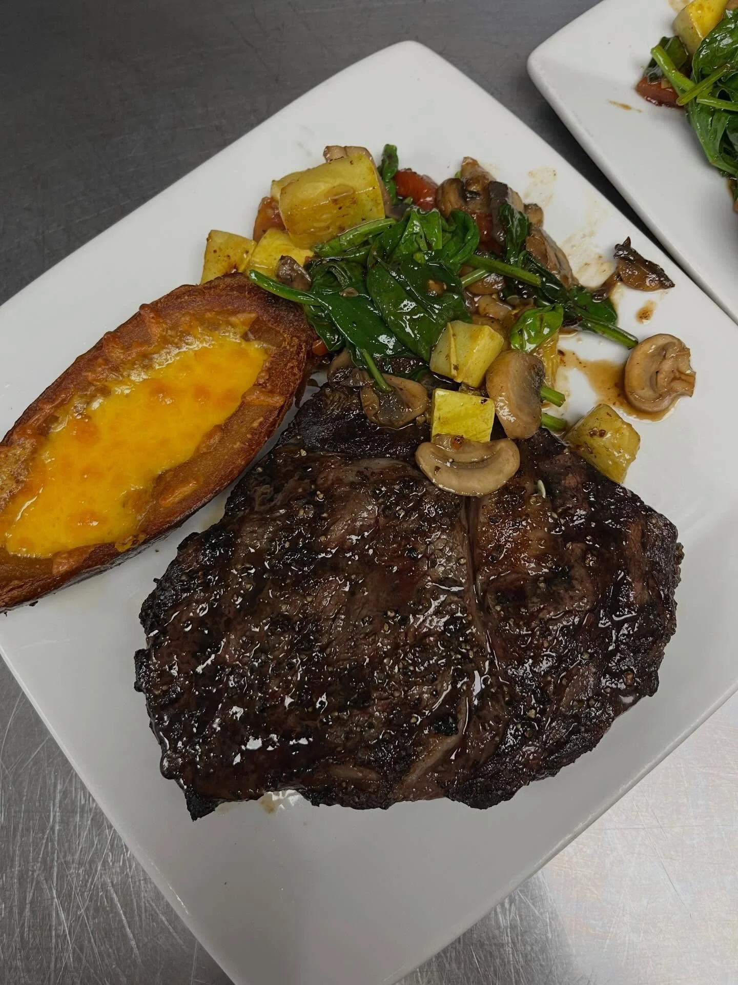 A white plate with a grilled steak topped with a brown sauce, roasted potatoes, sautéed mushrooms, sautéed spinach, and a piece of cheesy garlic bread.