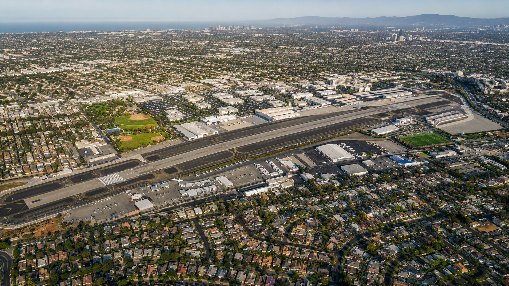 Aerial view of a large airport with runways and taxiways surrounded by residential neighborhoods, commercial buildings, and green parks, with mountains and city skyline in the background.