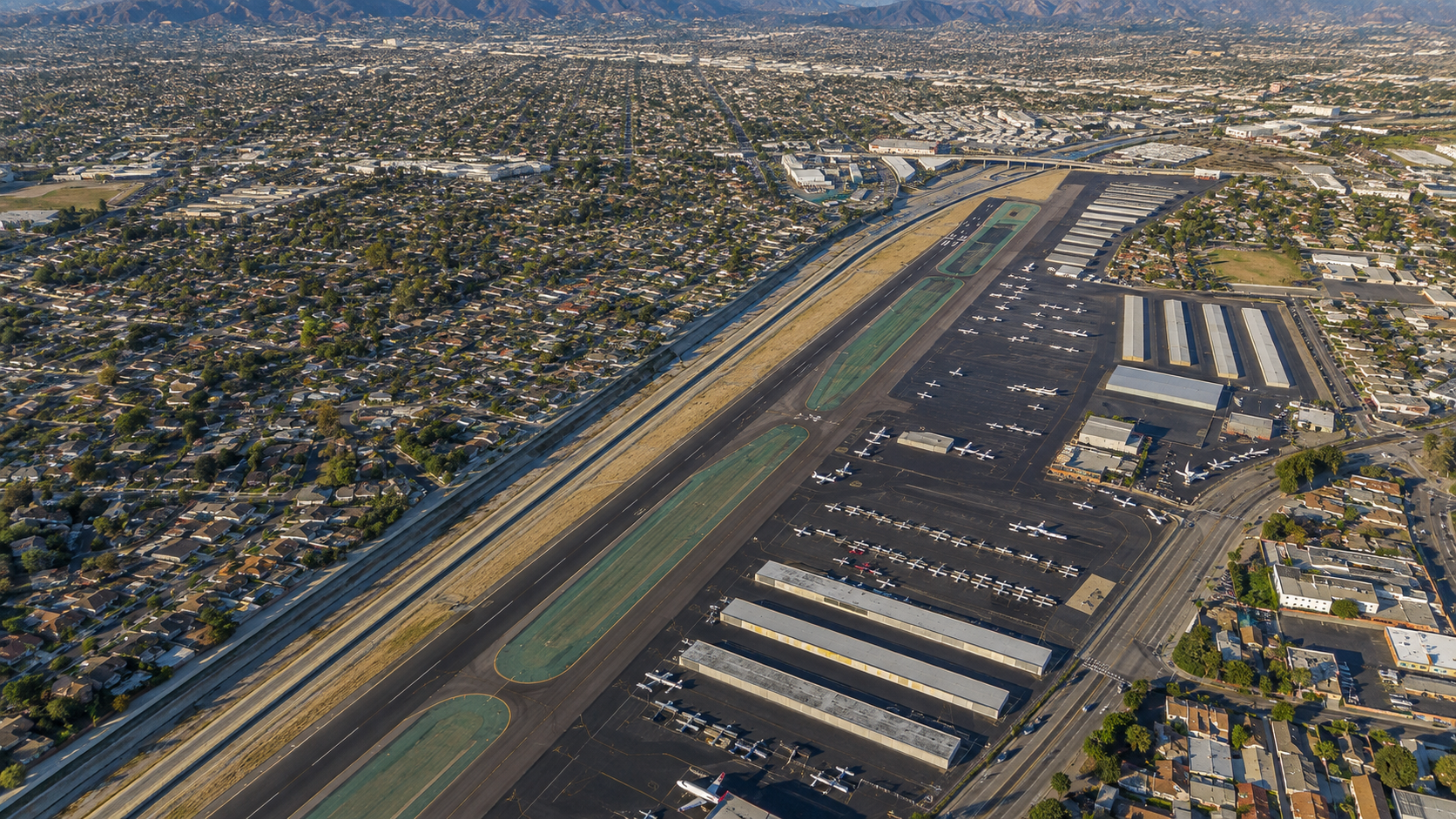An aerial view of an airport with planes parked on the tarmac, runways, surrounding commercial buildings, and a densely populated residential area with houses and streets.