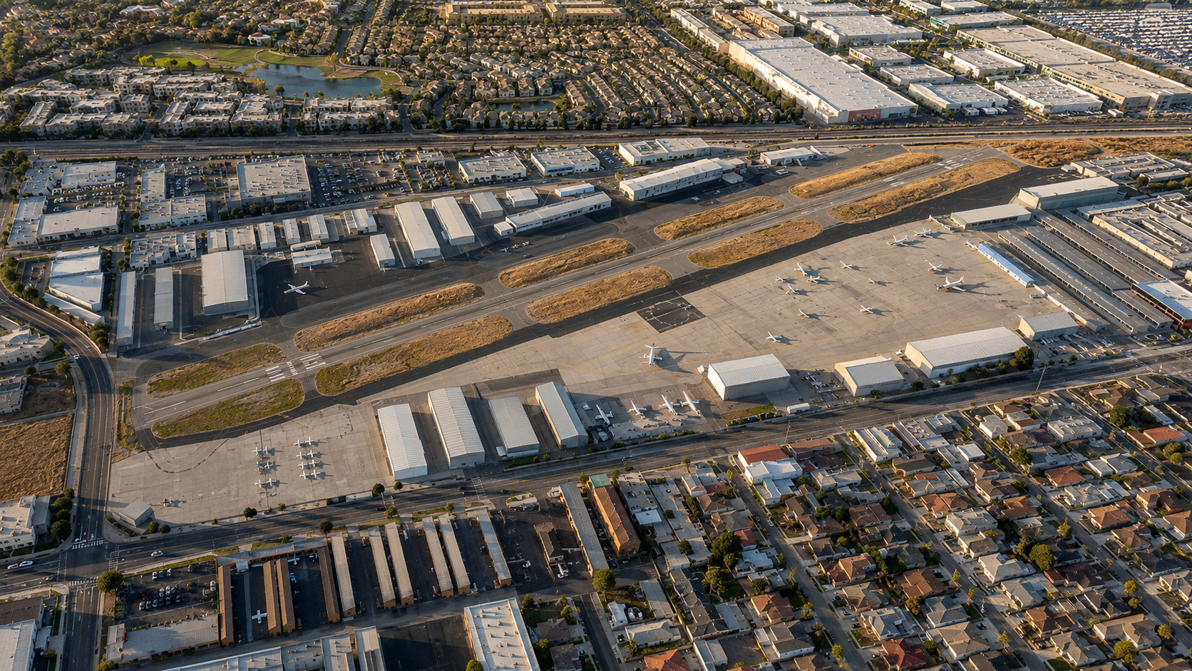Aerial view of an airport with a runway, several jets parked on the tarmac, and surrounding buildings, roads, and residential neighborhoods.