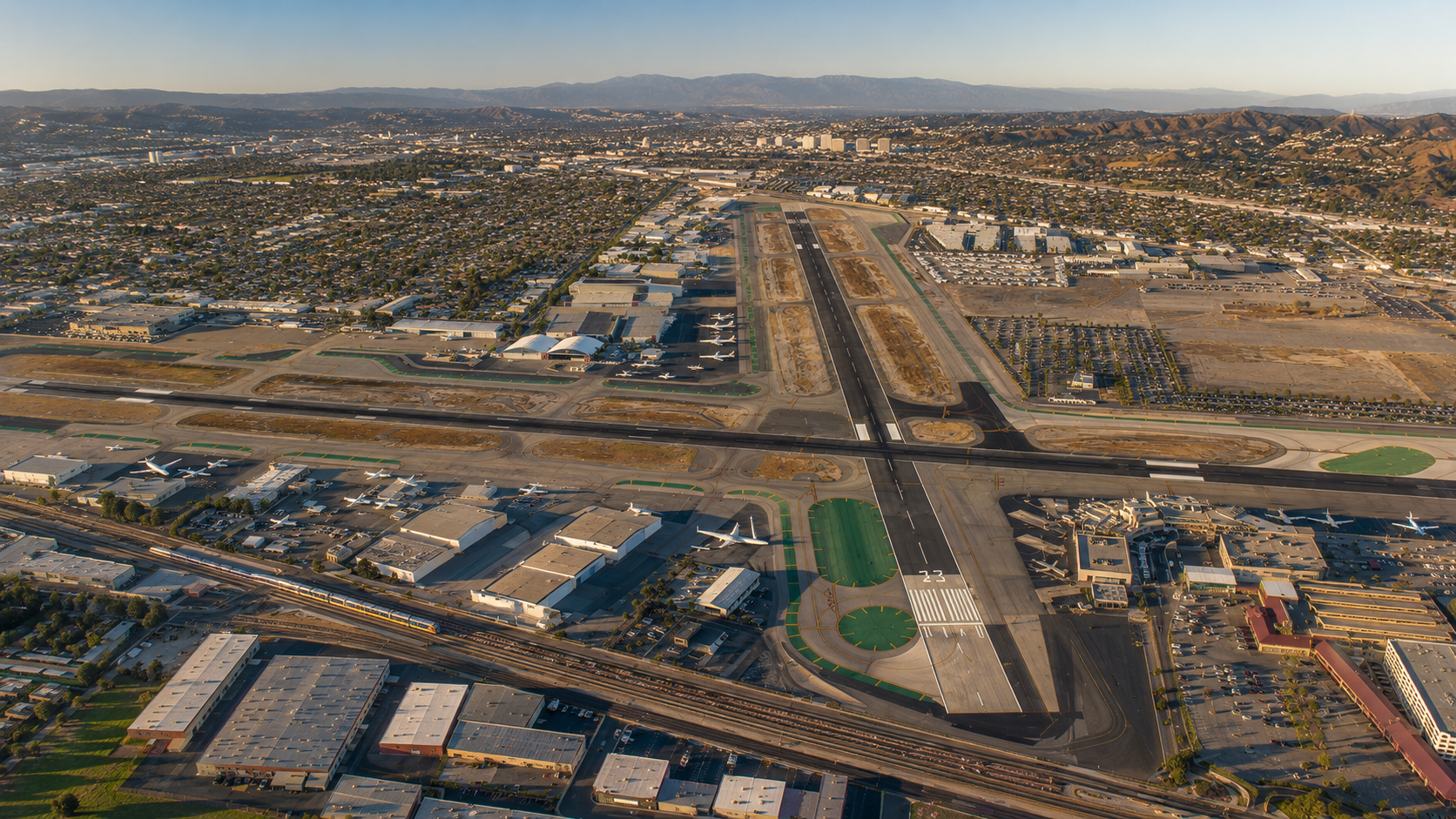 Aerial view of an airport runway during daytime, with airplanes parked near terminals, surrounding industrial buildings, highways, and a cityscape with hills in the background.