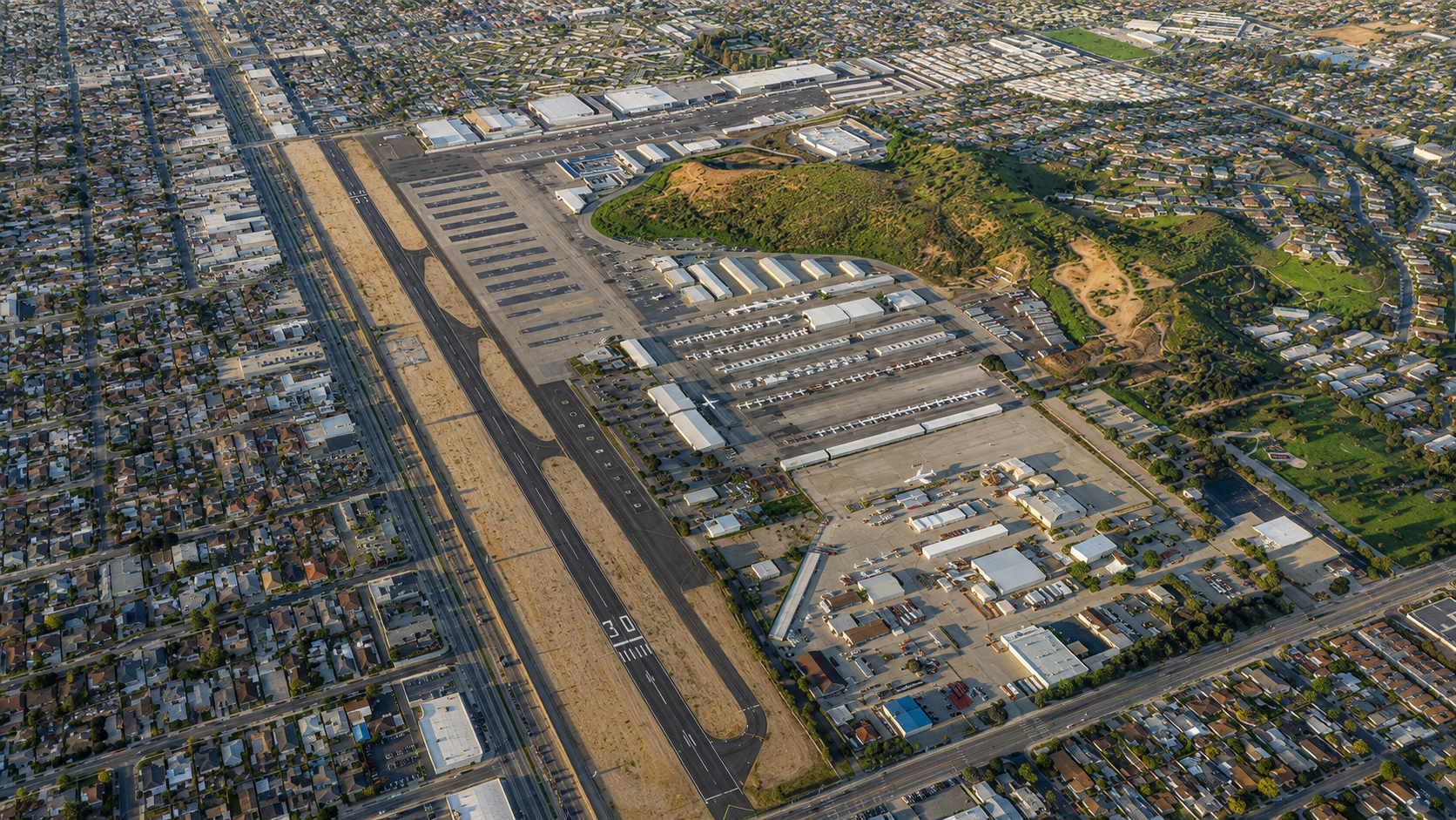 Aerial view of an airport with runways, taxiways, large hangars, aircraft parked at terminal gates, surrounding urban area, residential neighborhoods, and green park areas.