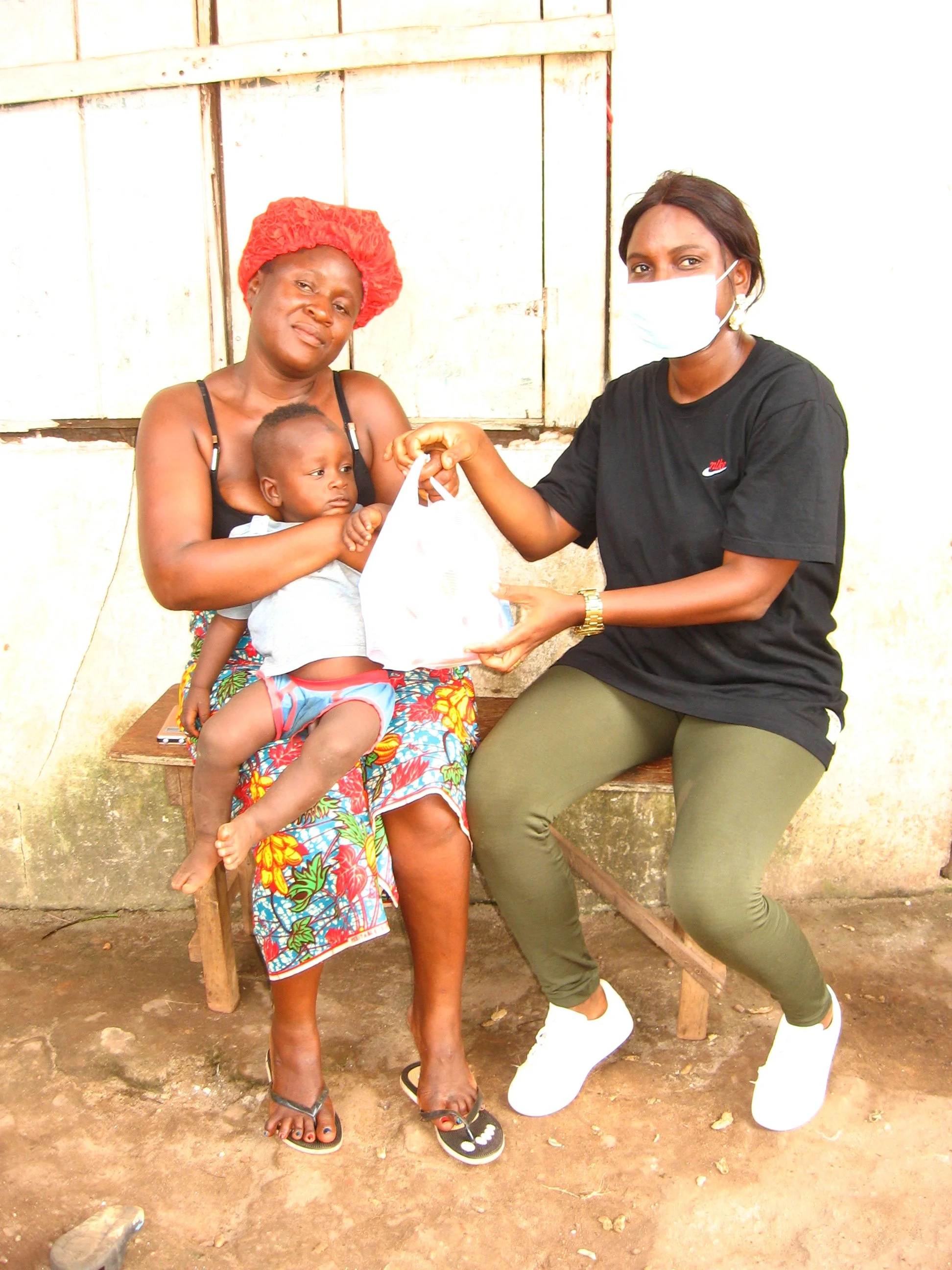 A woman holding a baby and an exchange of a paper bag between her and a second woman wearing a face mask. They are sitting outside against a wooden wall.
