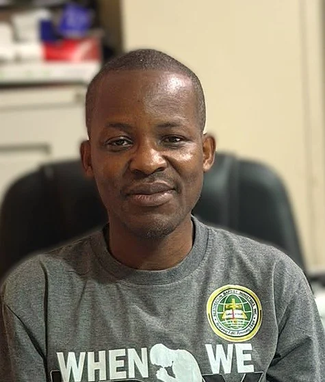 A man sitting in an office, wearing a gray T-shirt with a logo on the chest, in front of a blurred background with office supplies.