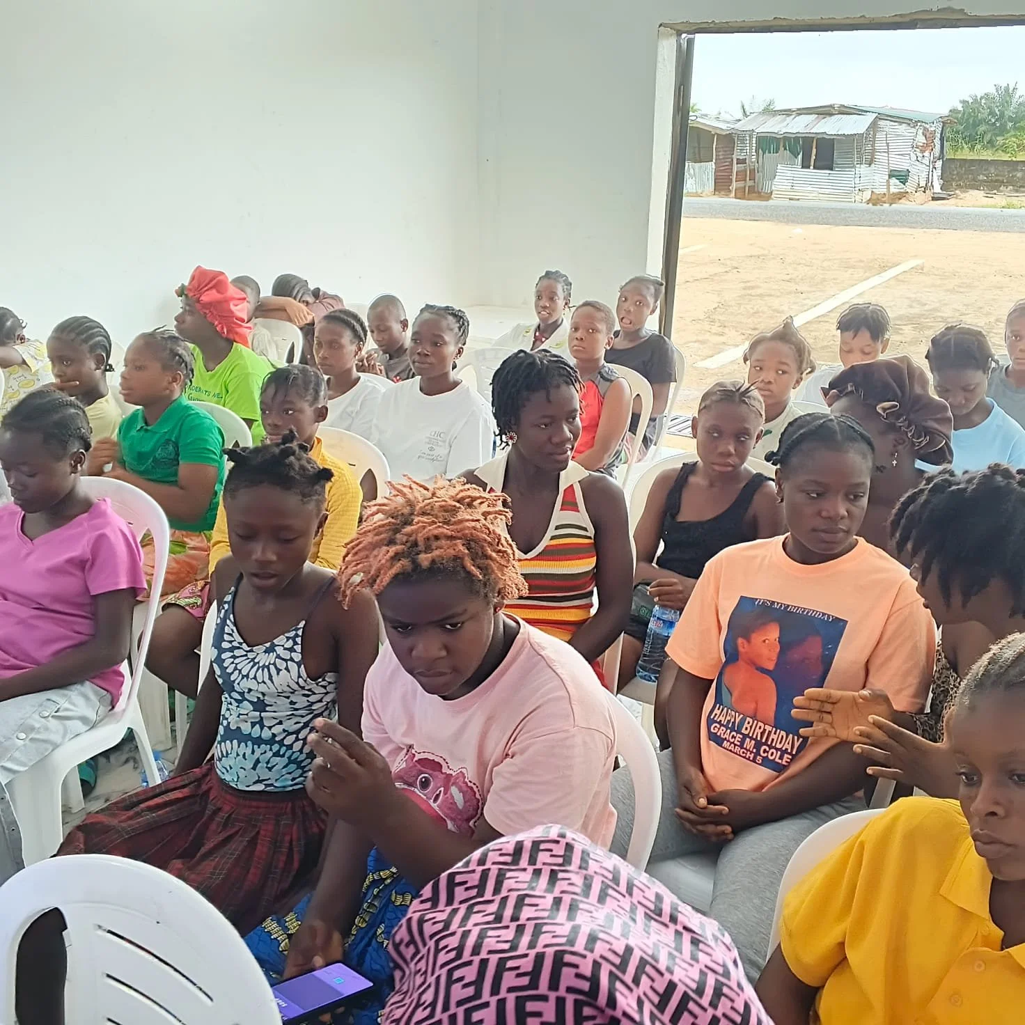 Group of children and young teens sitting indoors, some looking at phones, with a partially open door showing an outdoor area with dirt ground and makeshift structures.