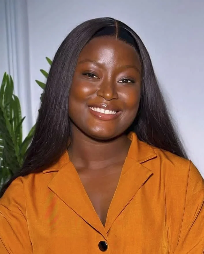 A smiling woman with long straight hair, wearing an orange blazer, standing in front of a plant and a neutral background.