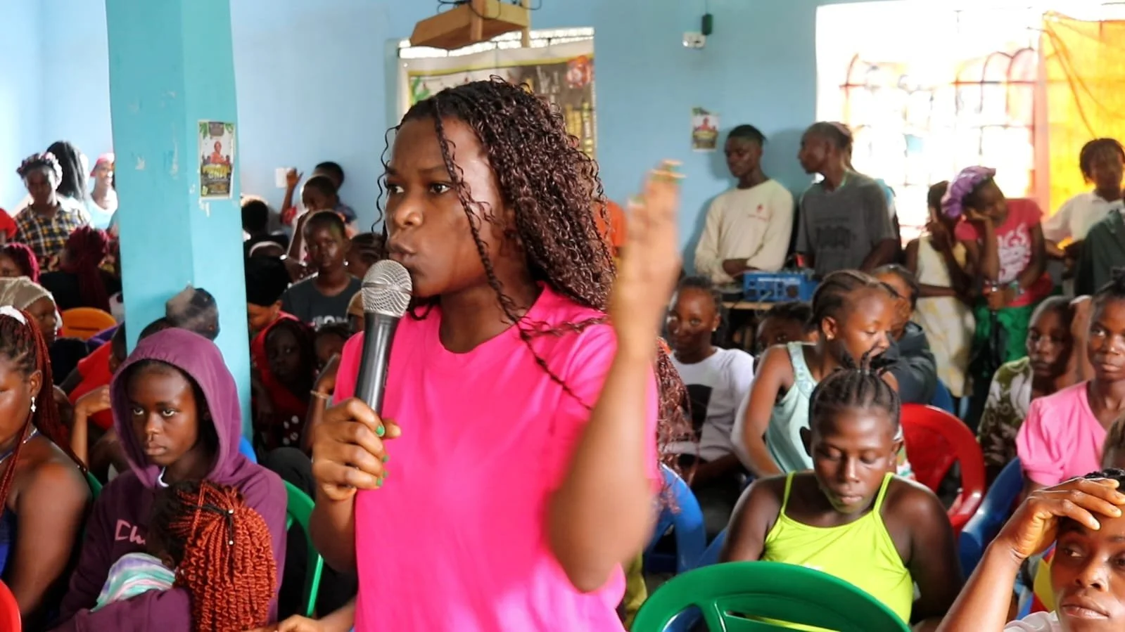 A woman in a pink shirt speaks into a microphone during a gathering of people in a blue-walled room, with many women and girls seated and standing around her.
