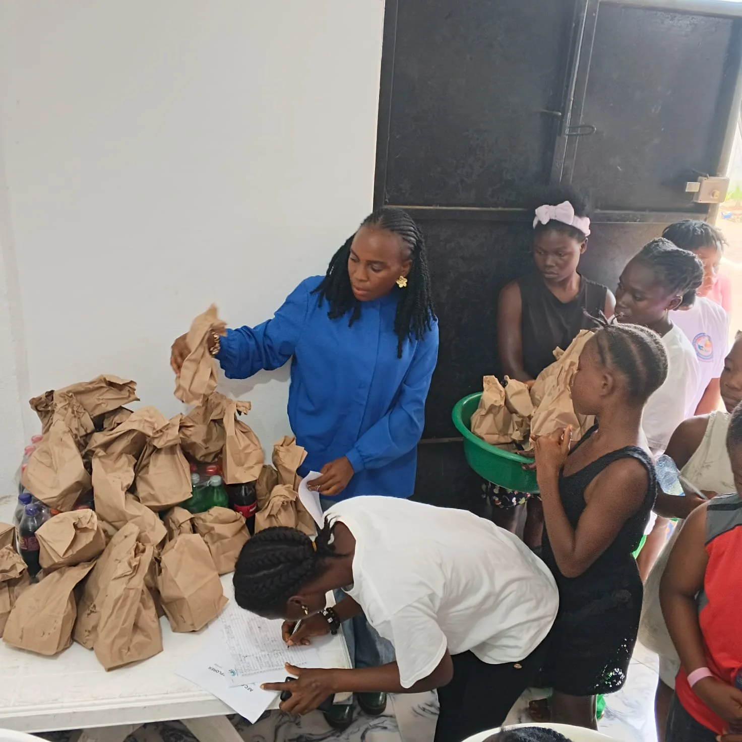A group of women and girls in line at a food distribution table with brown paper bags and bottled drinks, in an indoor setting.