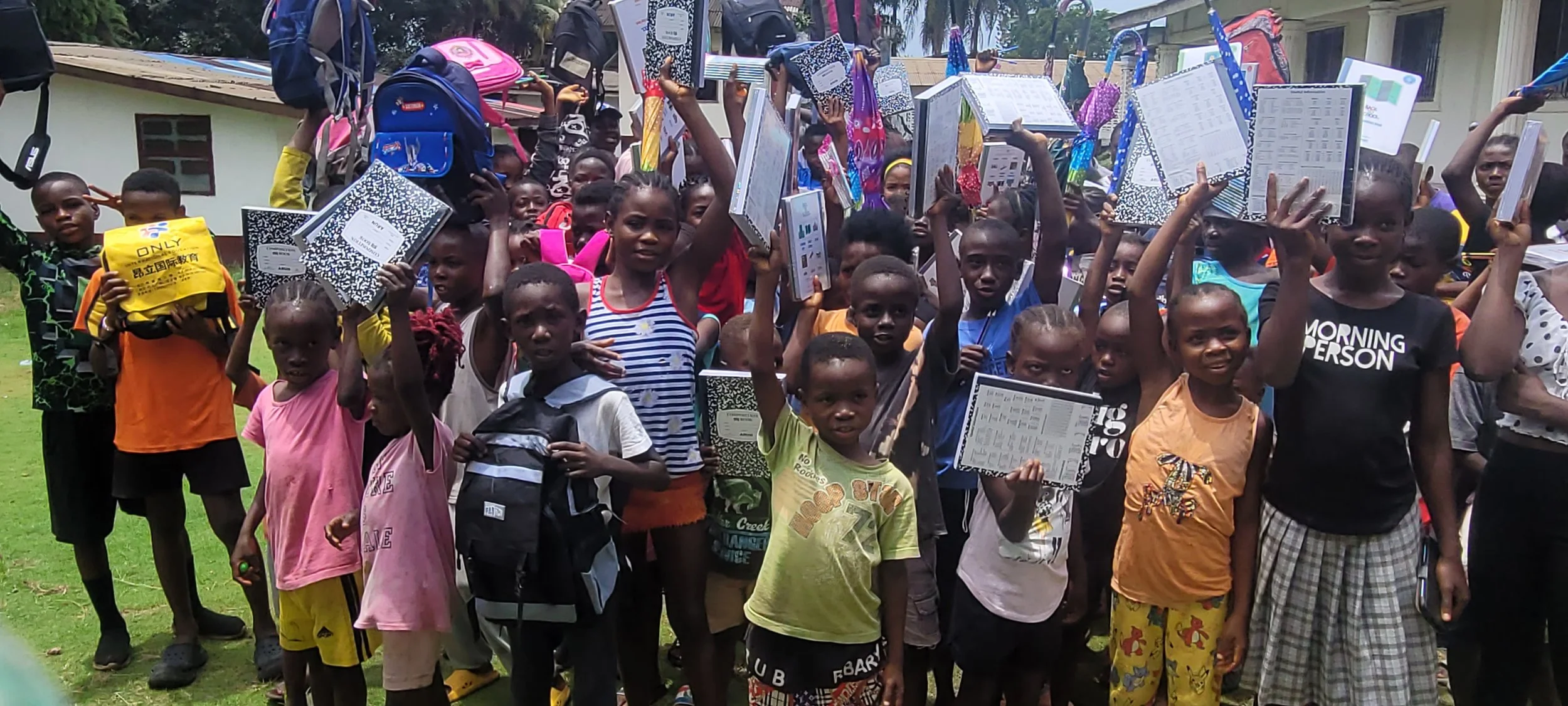 Group of children and teenagers outdoors, holding notebooks and backpacks, with some raising their hands. They appear happy and excited, with buildings and trees in the background.
