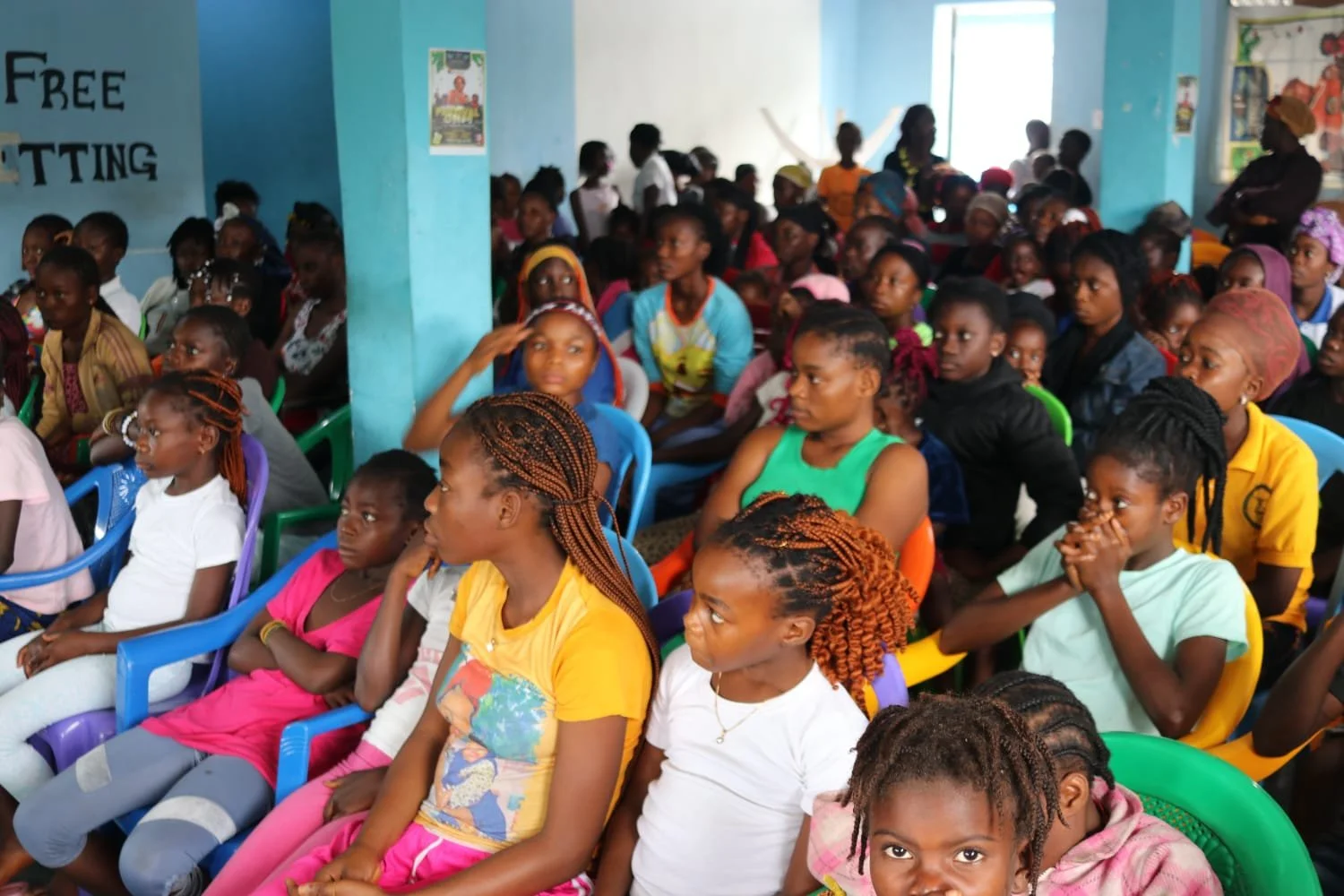 A large group of children and young girls sitting on colorful chairs indoors, attending an event or gathering, with a blue wall and posters in the background.