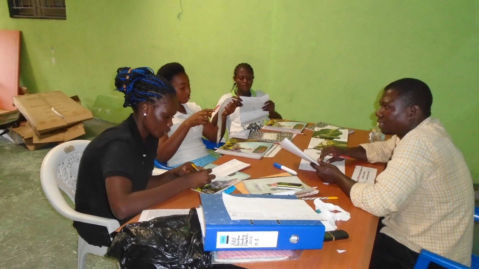 Five people sit around a table reviewing documents and brochures in a room with green walls. Three women and one man are engaged in discussion, with magazines and papers spread across the table.