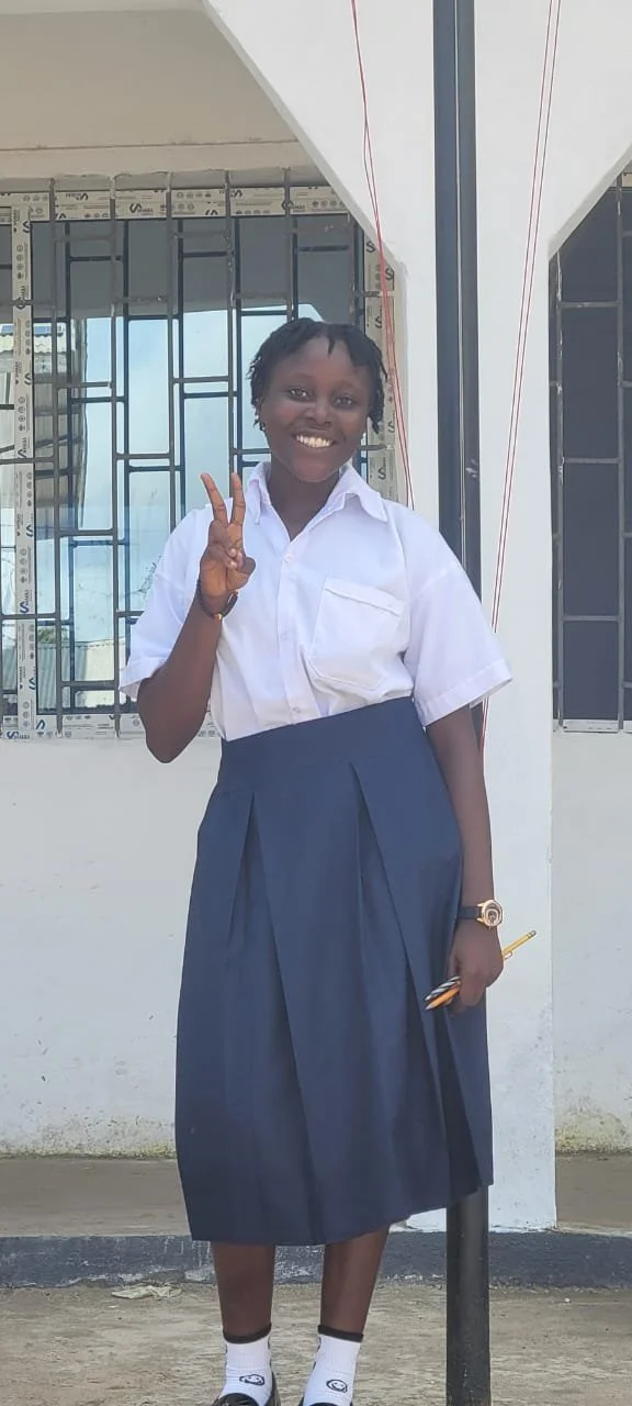 A girl in school uniform standing outside, smiling and making a peace sign with her right hand, holding a smartphone in her left hand.