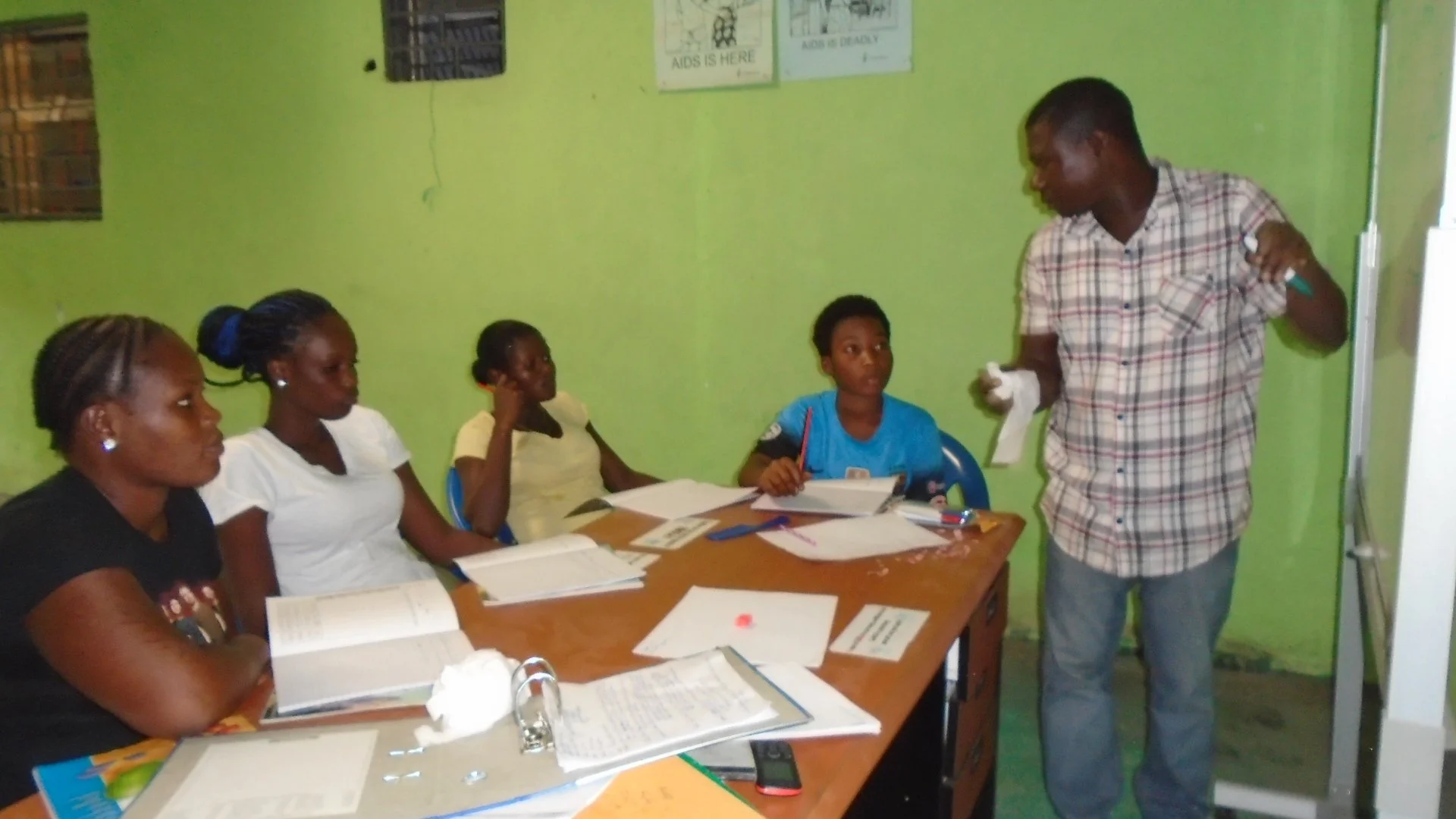 A classroom scene with five people sitting at a table and one standing. The instructor appears to be teaching the students, who are listening attentively. The classroom has green walls, and there are posters on the wall. The table is cluttered with books, papers, and writing materials.