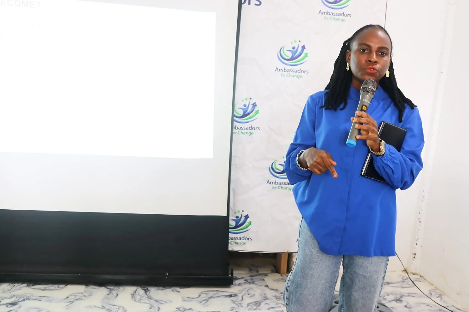A woman wearing a blue jacket holding a microphone and a notebook, standing in front of a white backdrop with the title "Ambassadors for Change" and a logo.