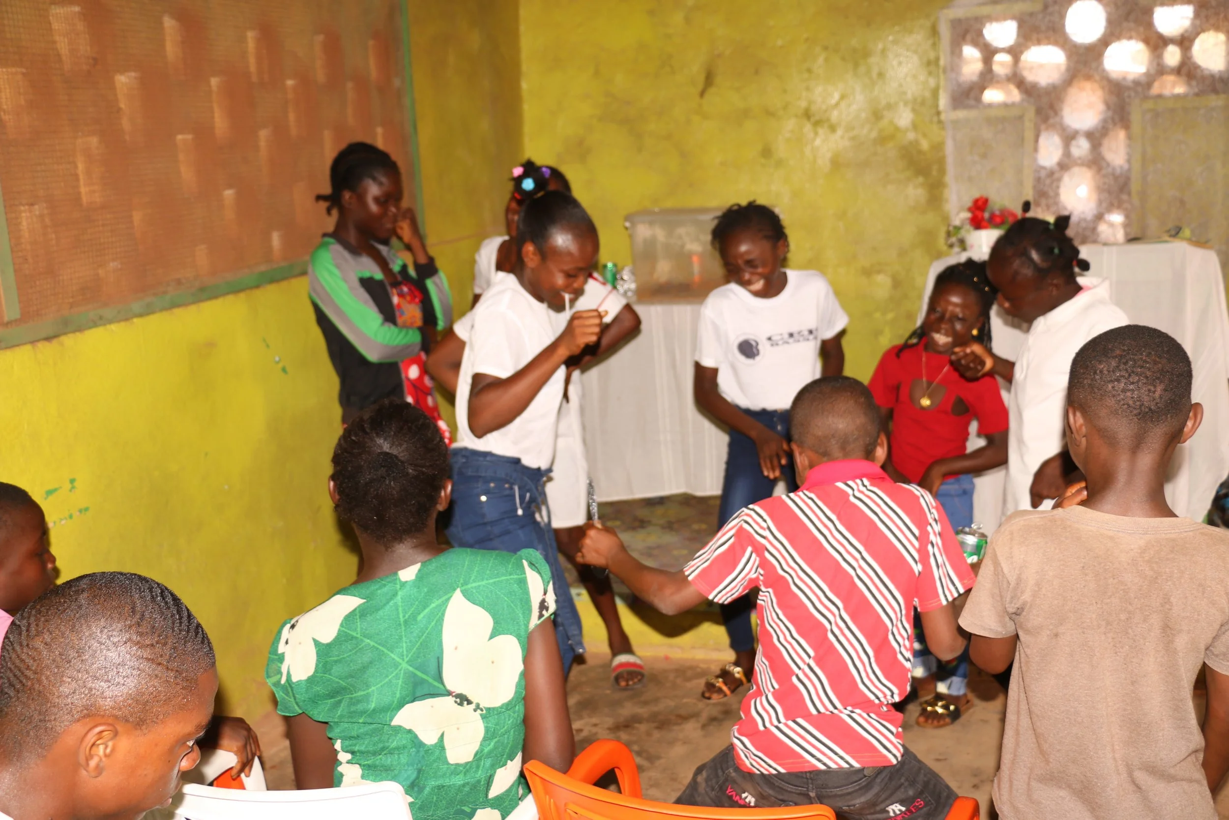 Group of children and women with dark skin smiling and playing in a room with yellow walls and green and brick accents.
