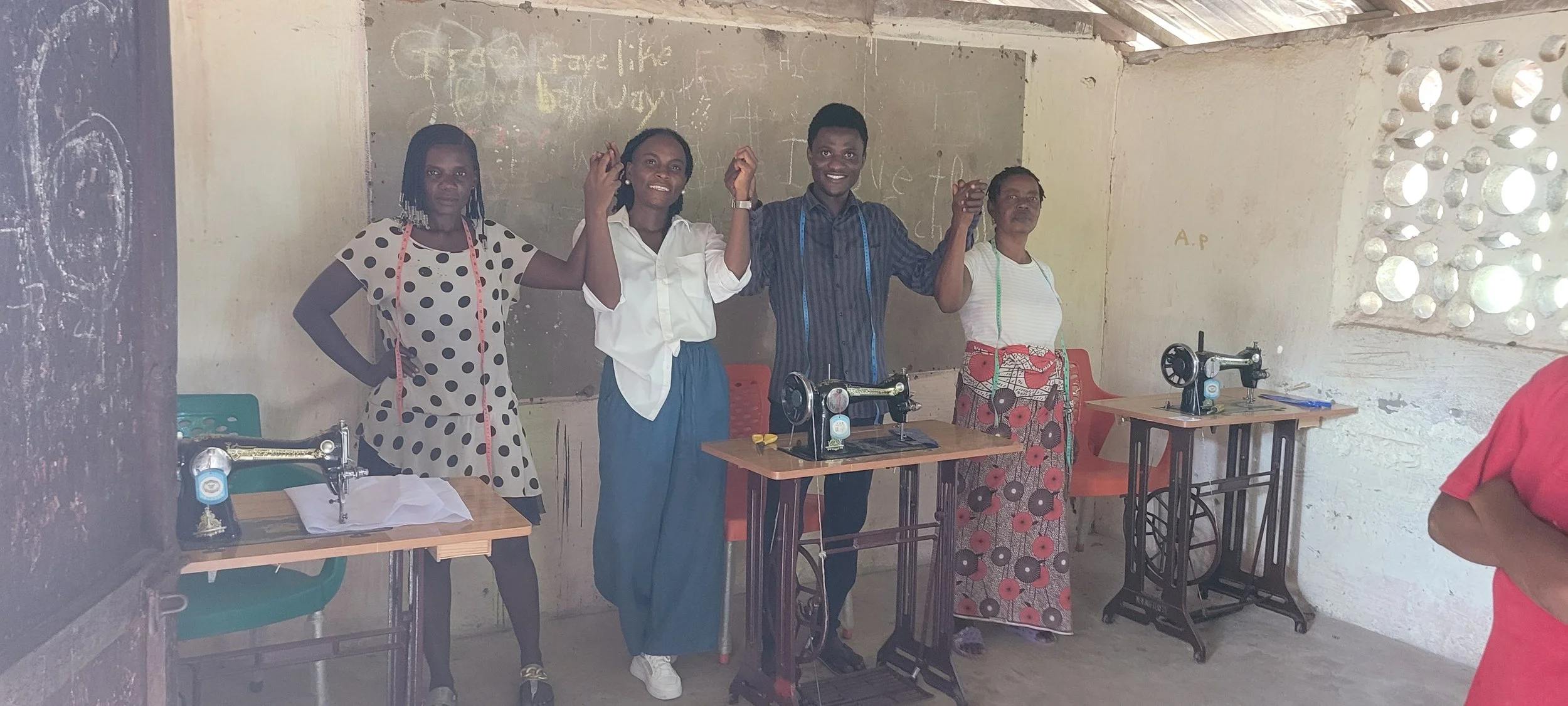 Four women standing in a classroom with sewing machines, holding hands and smiling, in front of a blackboard and a wall with holes allowing light to pass through.