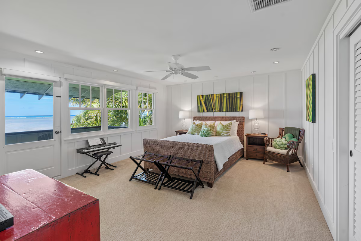 Bright bedroom with white paneled walls, large windows showing a view of palm trees and the beach, a bed with pillows and green decor, nightstands with lamps, a wicker chair, and a small keyboard near the window.