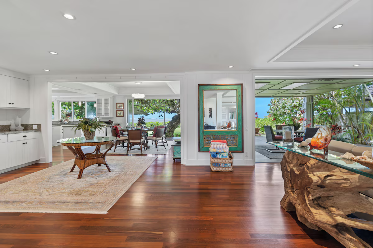 Interior view of a spacious, bright living area with wooden floors, white walls, and large windows showing outdoor greenery and ocean. The room features a round wooden table with a flower arrangement, a rug, and a decorative mirror on the wall. There