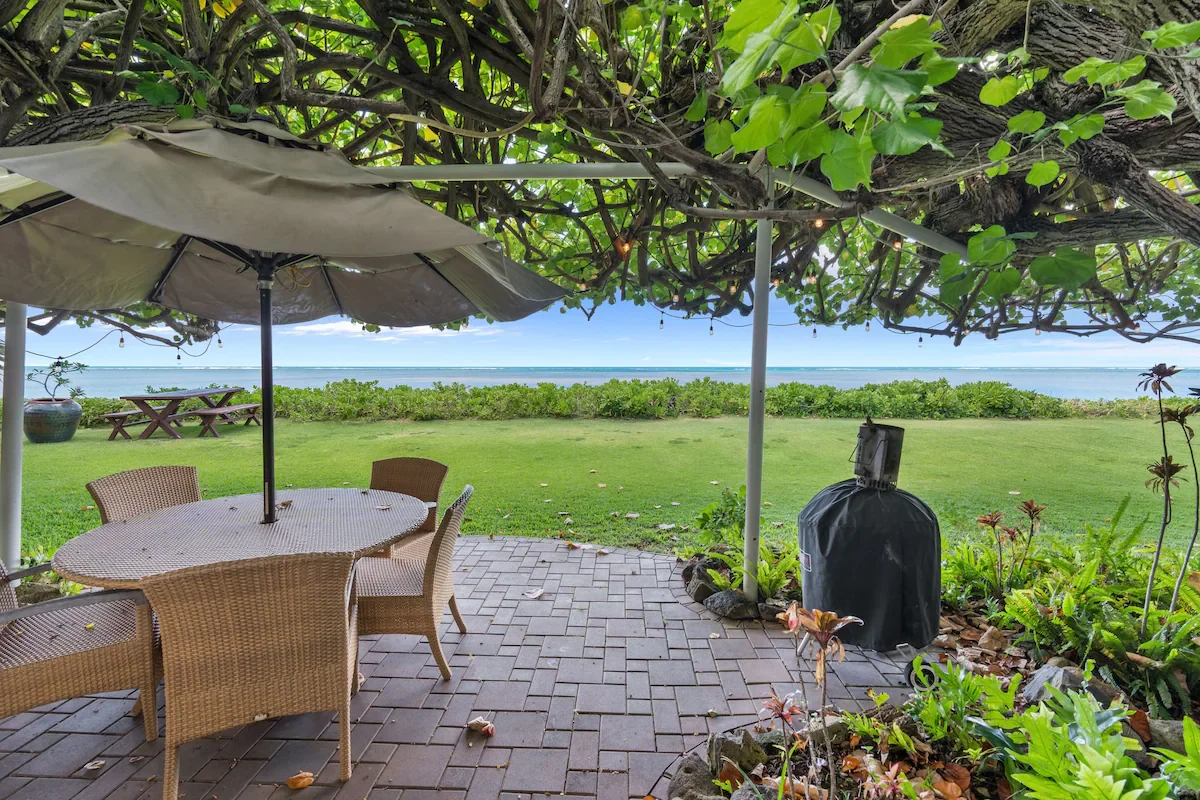 A patio with a wicker dining set and an umbrella, overlooking a grassy yard with a view of the ocean, under a large leafy tree.