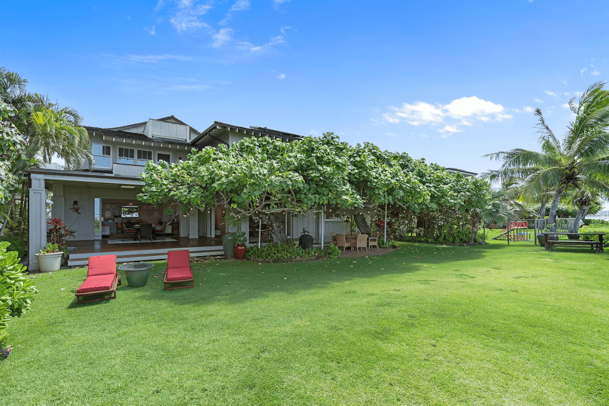 A backyard with green grass, two red lounge chairs, and tropical trees including palm trees. There is a patio with outdoor furniture and a house in the background under a blue sky.