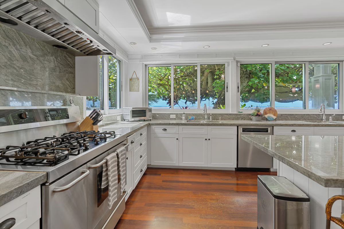 Bright kitchen with white cabinets, granite countertops, stainless steel appliances, and a large window showing green trees outside.