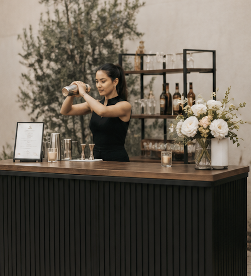 A professional bartender shaking a cocktail behind a bar with floral decorations and a background mural of trees.