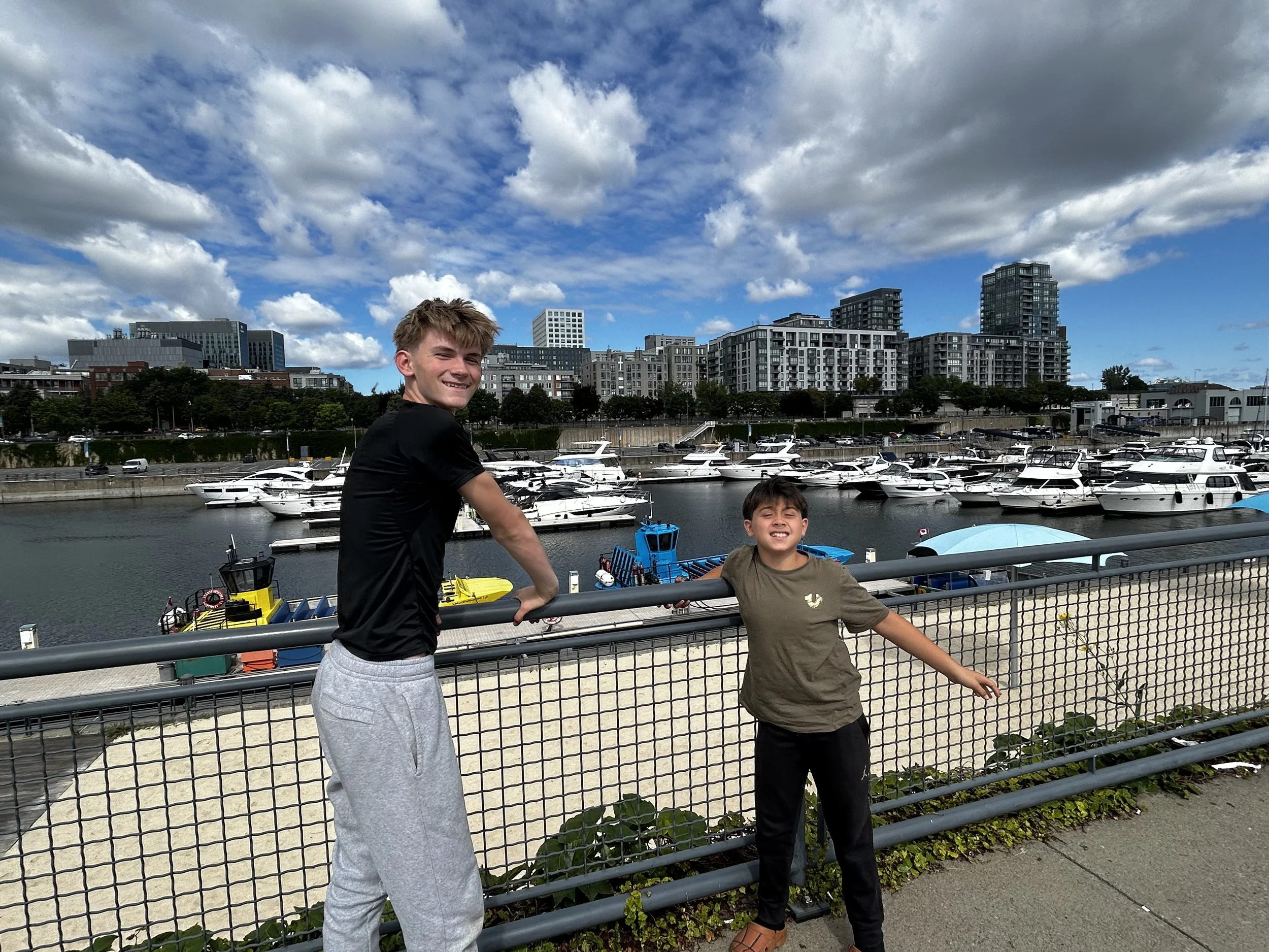 Two boys standing by a pier with boats, high-rise buildings in the background, and a partly cloudy sky.