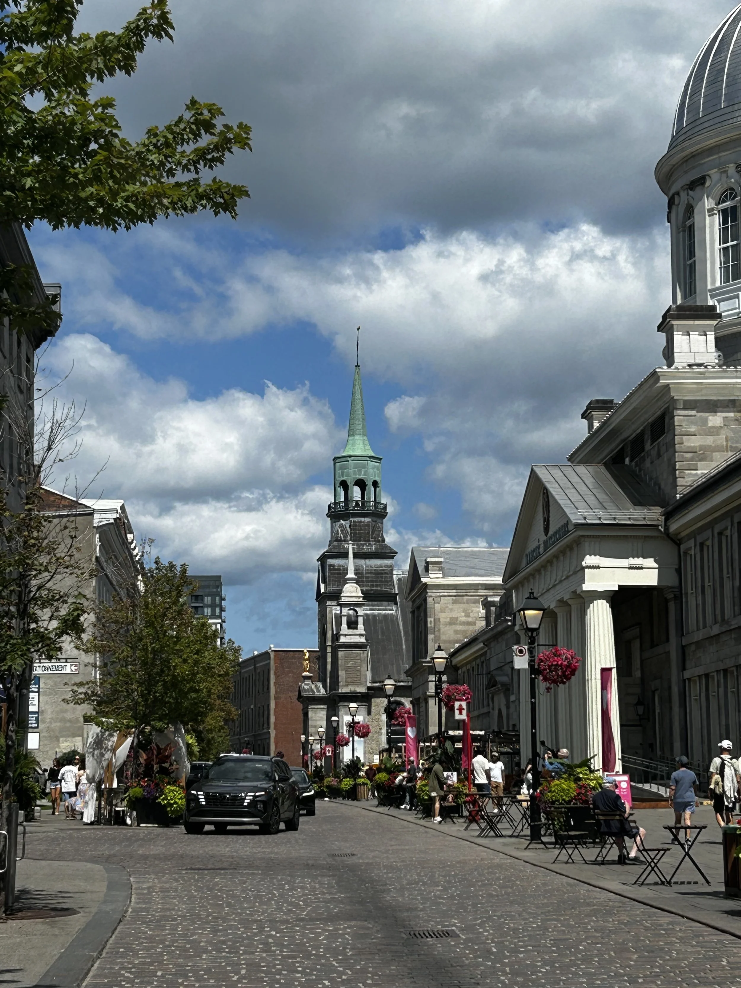 A busy city street with historic buildings, outdoor seating, and people walking. A church steeple is visible against a partly cloudy sky.