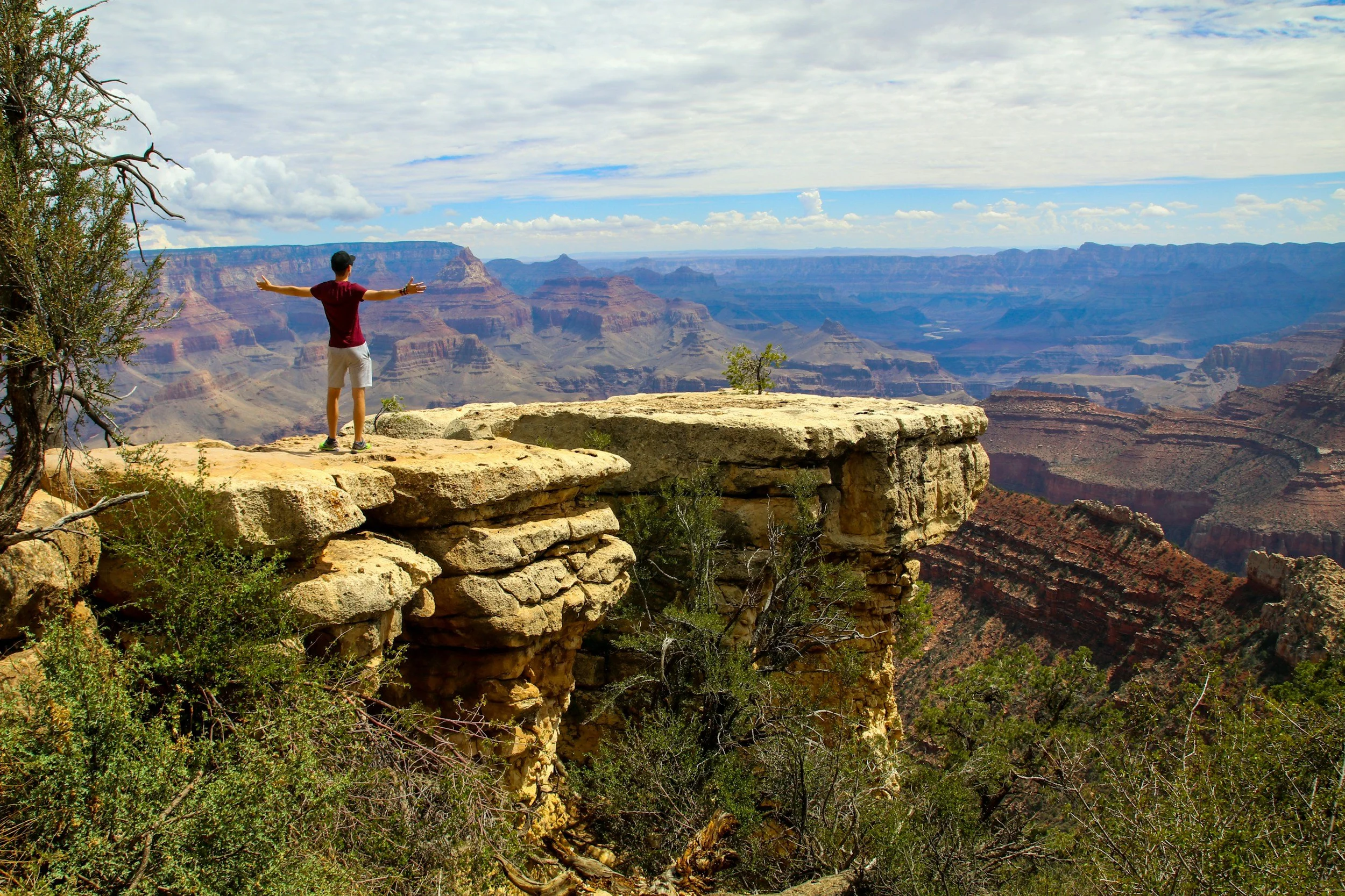 A person standing on a rocky ledge with arms outstretched overlooking the Grand Canyon on a partly cloudy day.