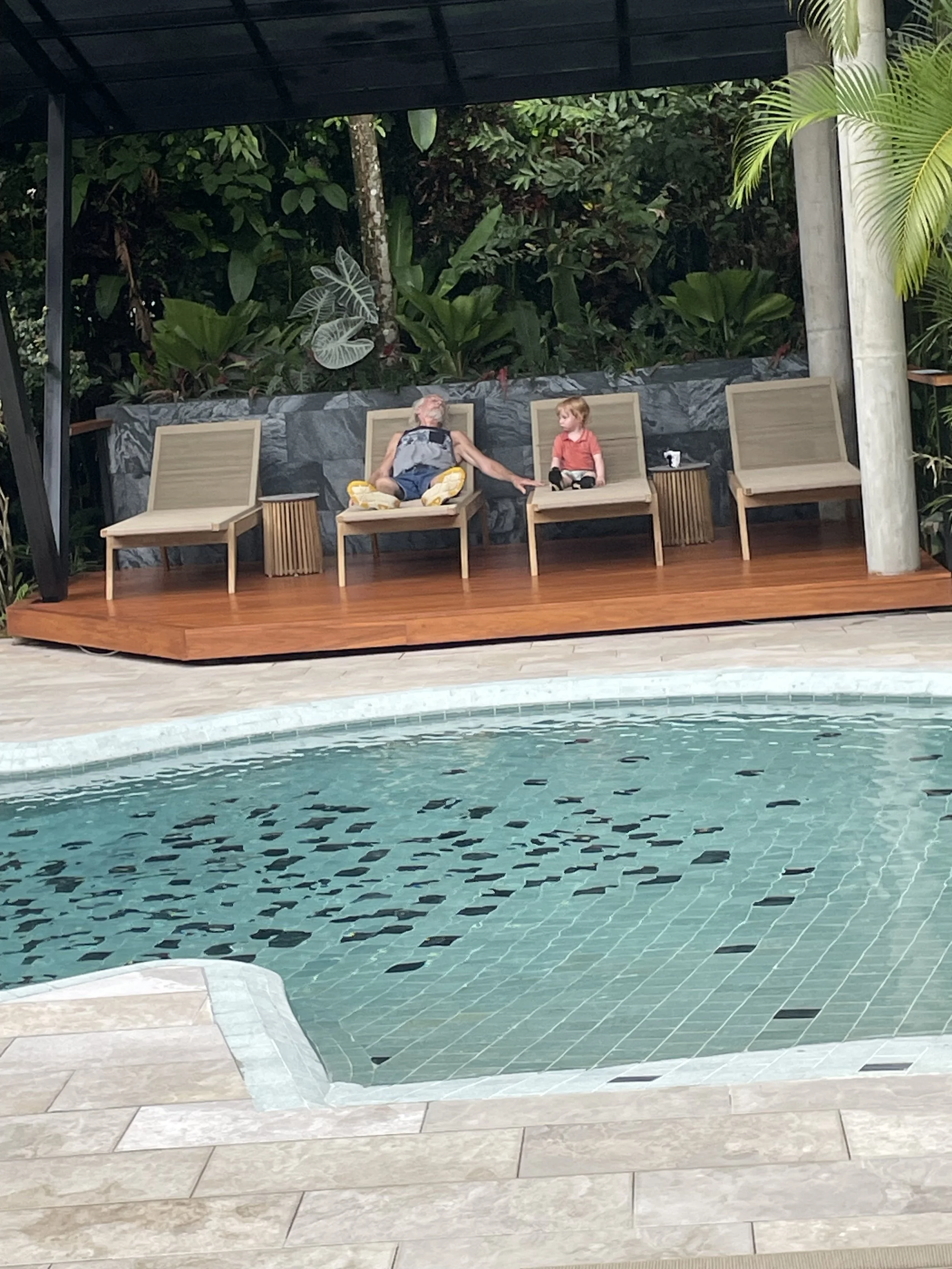 A swimming pool with clear water and black mosaic tiles, surrounded by light-colored stone tiles, and a wooden deck area with seating. Two people are sitting on chairs by a stone wall, one elderly man and a young boy, with lush greenery in the background.