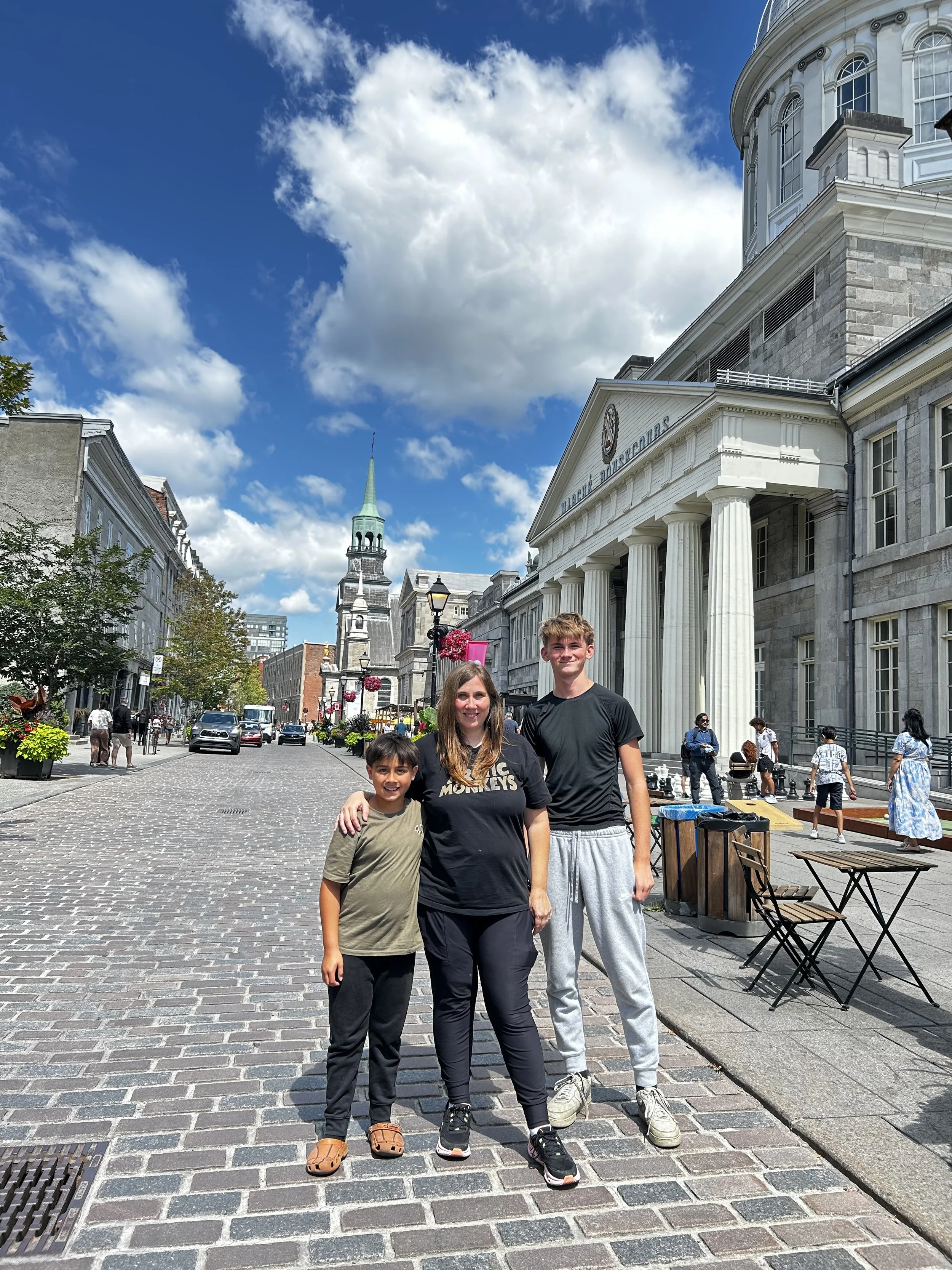 A group of three people, a woman and two young boys, standing on a cobblestone street in front of a historic building with large columns and a sign that reads "Library Bounded" in an urban area with other pedestrians, trees, and buildings visible under a blue sky with scattered clouds.