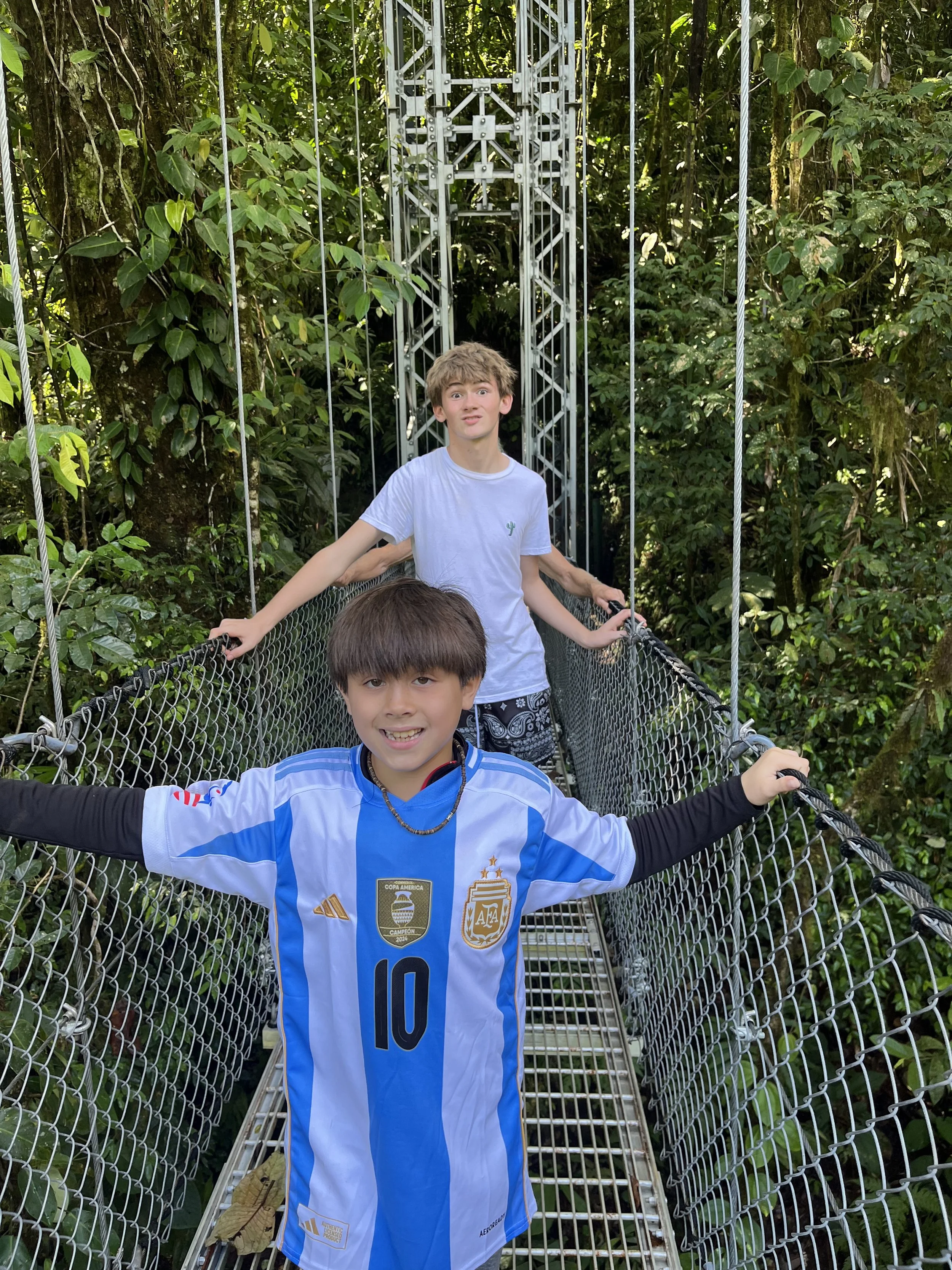 Two young boys standing on a suspension bridge surrounded by lush green tropical forest. The boy in the foreground is wearing a blue and white soccer jersey with the number 10, and the boy in the background is wearing a white T-shirt with a cactus logo. They are holding onto the railing of the bridge.