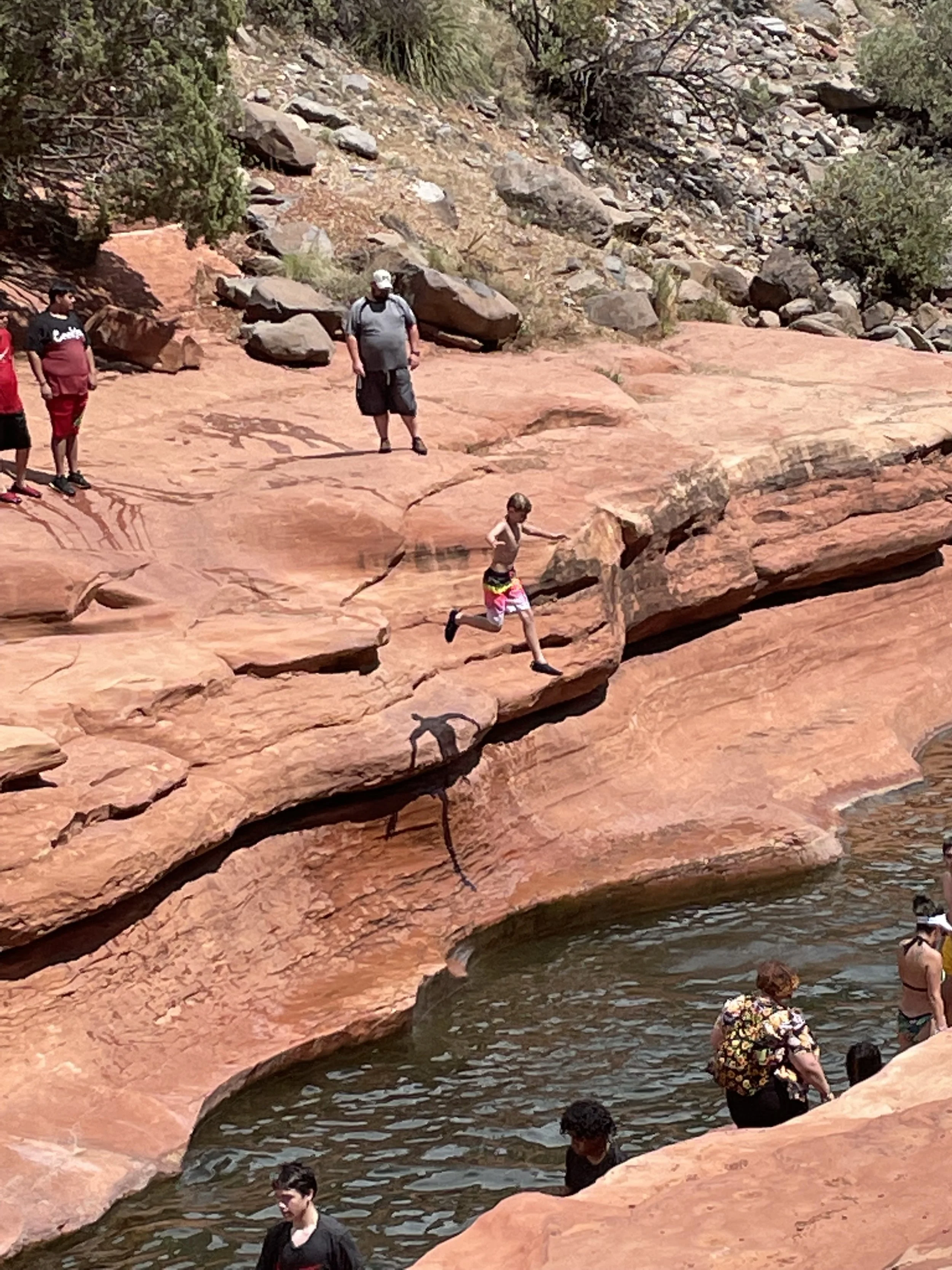 People enjoying a rocky river with red and brown rocks, some swimming and others standing or running near the water’s edge, in a natural outdoor setting.