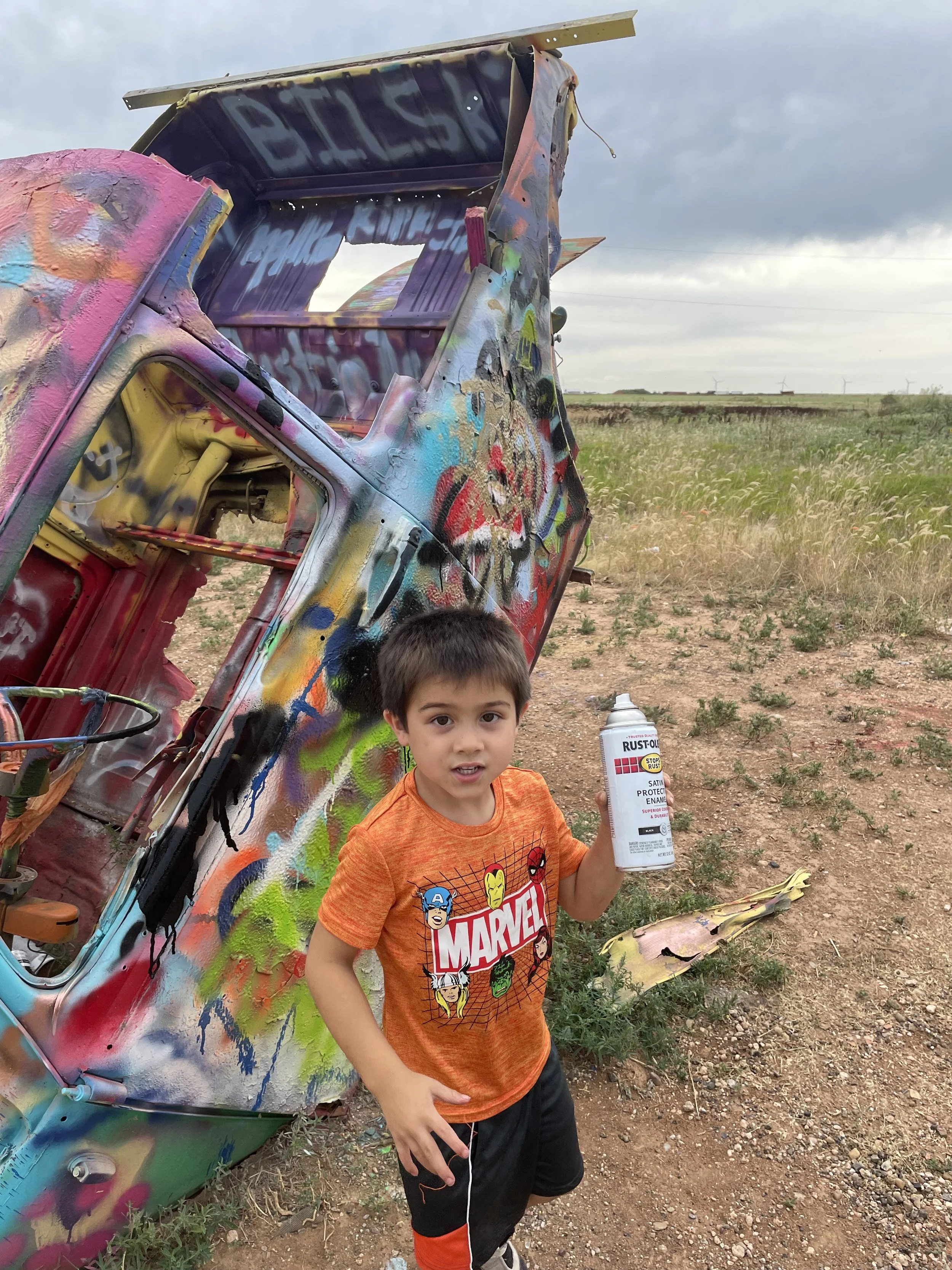 A young boy wearing a red Marvel T-shirt holding a can of rust-oleum spray paint while standing outdoors next to a colorful, graffiti-covered vehicle wreck in a field with wind turbines in the background.