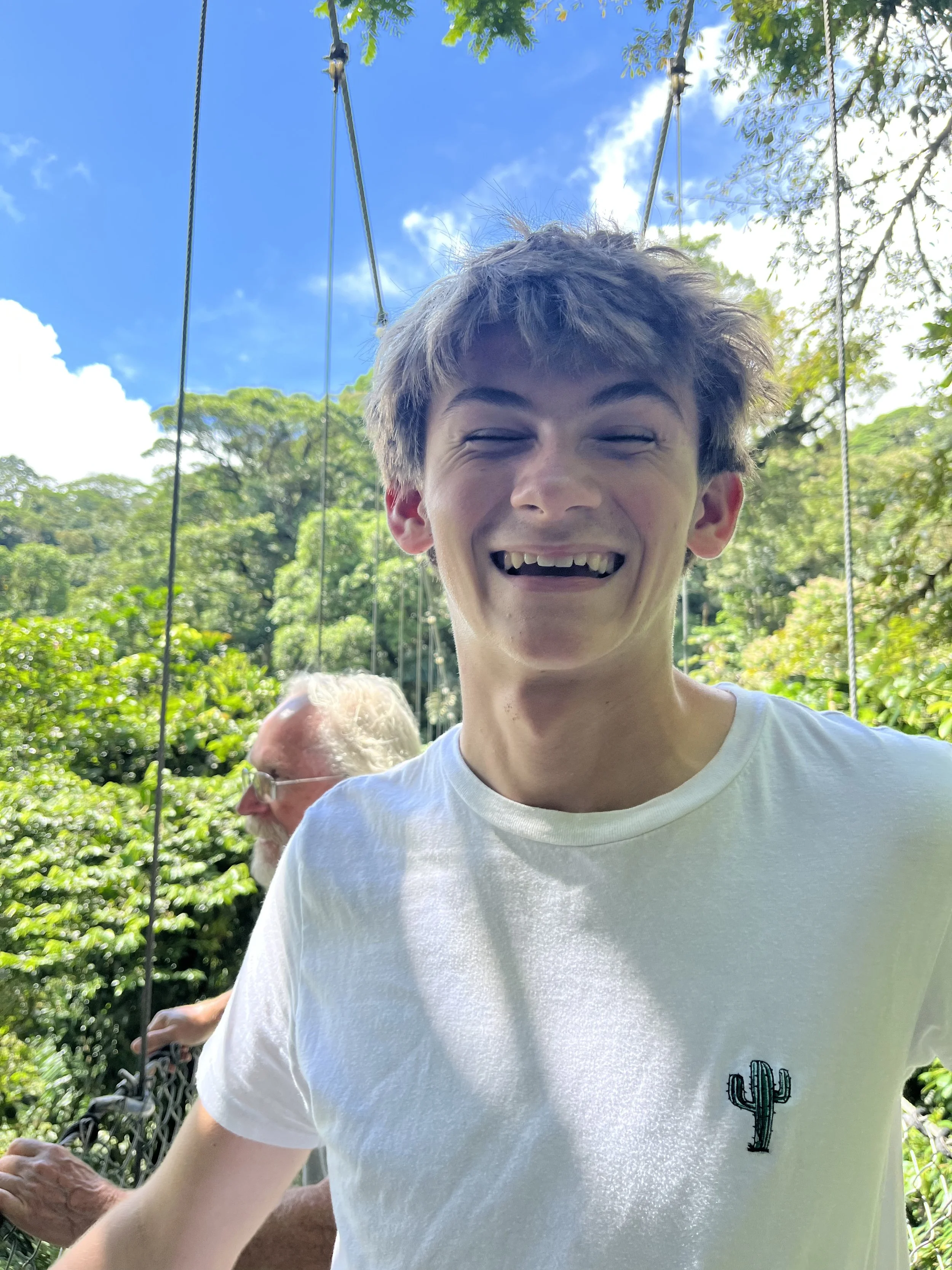 Smiling young man with short, blond hair and a visible gap in his front teeth, wearing a white t-shirt with a cactus embroidered on it, standing outdoors on a sunny day with blue sky and green trees in the background.