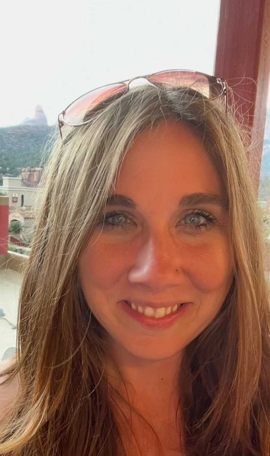 Close-up of a woman with long blonde hair and light-colored eyes, smiling at the camera. She has a small nose piercing and sunglasses resting on her head. The background shows a cityscape with buildings and a hill, possibly in a warm climate.