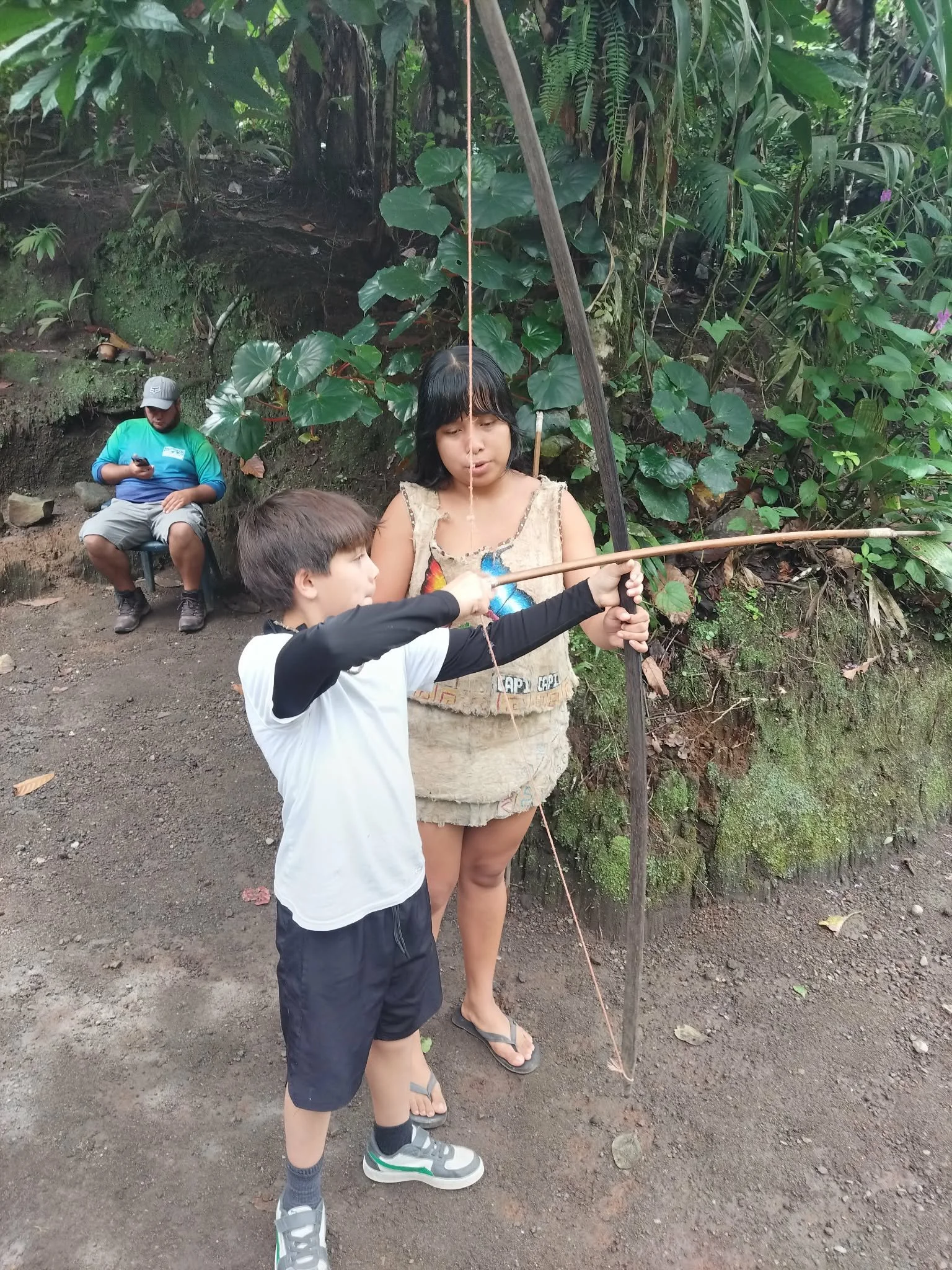 Two children are playing with a bow and arrow outdoors in a wooded area. One child is aiming the bow while the other helps. In the background, an adult is sitting on a chair, looking at a phone.