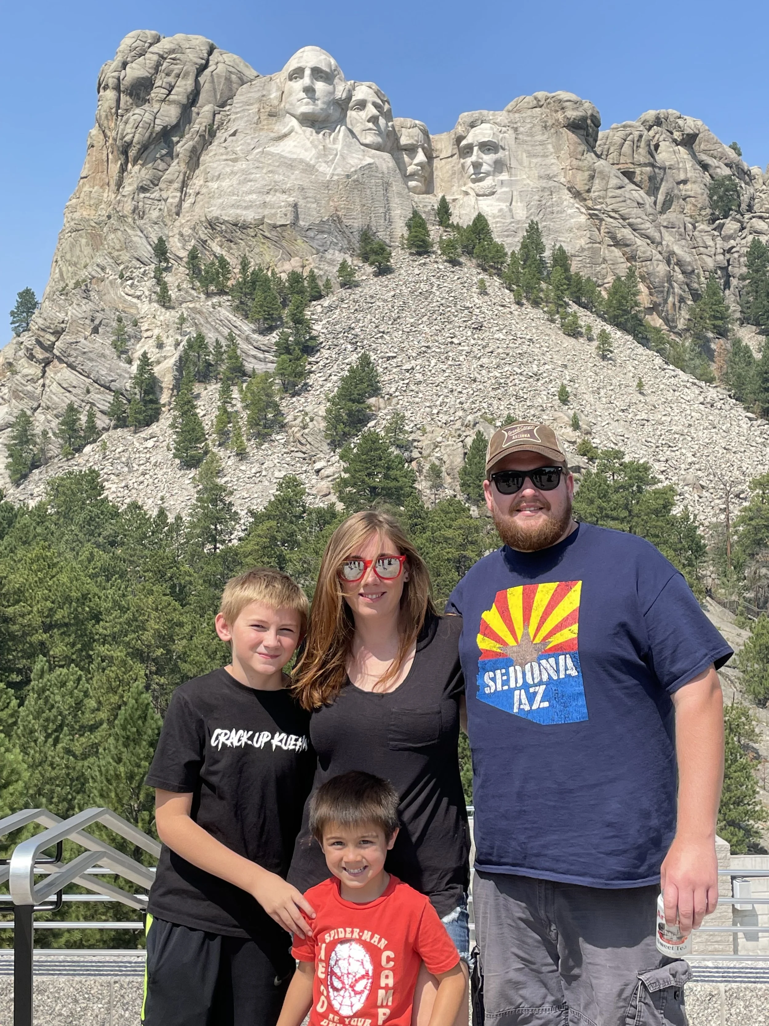 Group of four people — two children and two adults — smiling at the camera with Mount Rushmore in the background, outdoors on a sunny day.