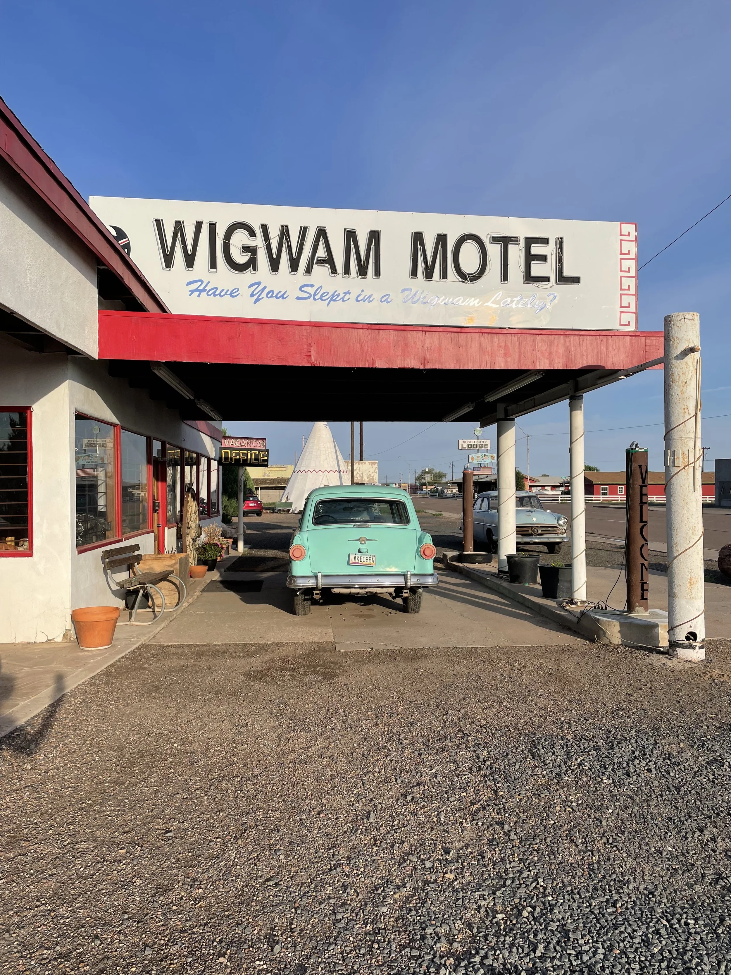 Small motel with a sign reading 'WIGWAM MOTEL' and a slogan 'Have You Slept in a Wigwam Lately?' above the parking area. There are vintage cars parked under the canopy, and the motel has a rustic, old-fashioned style with a teepee visible in the background.