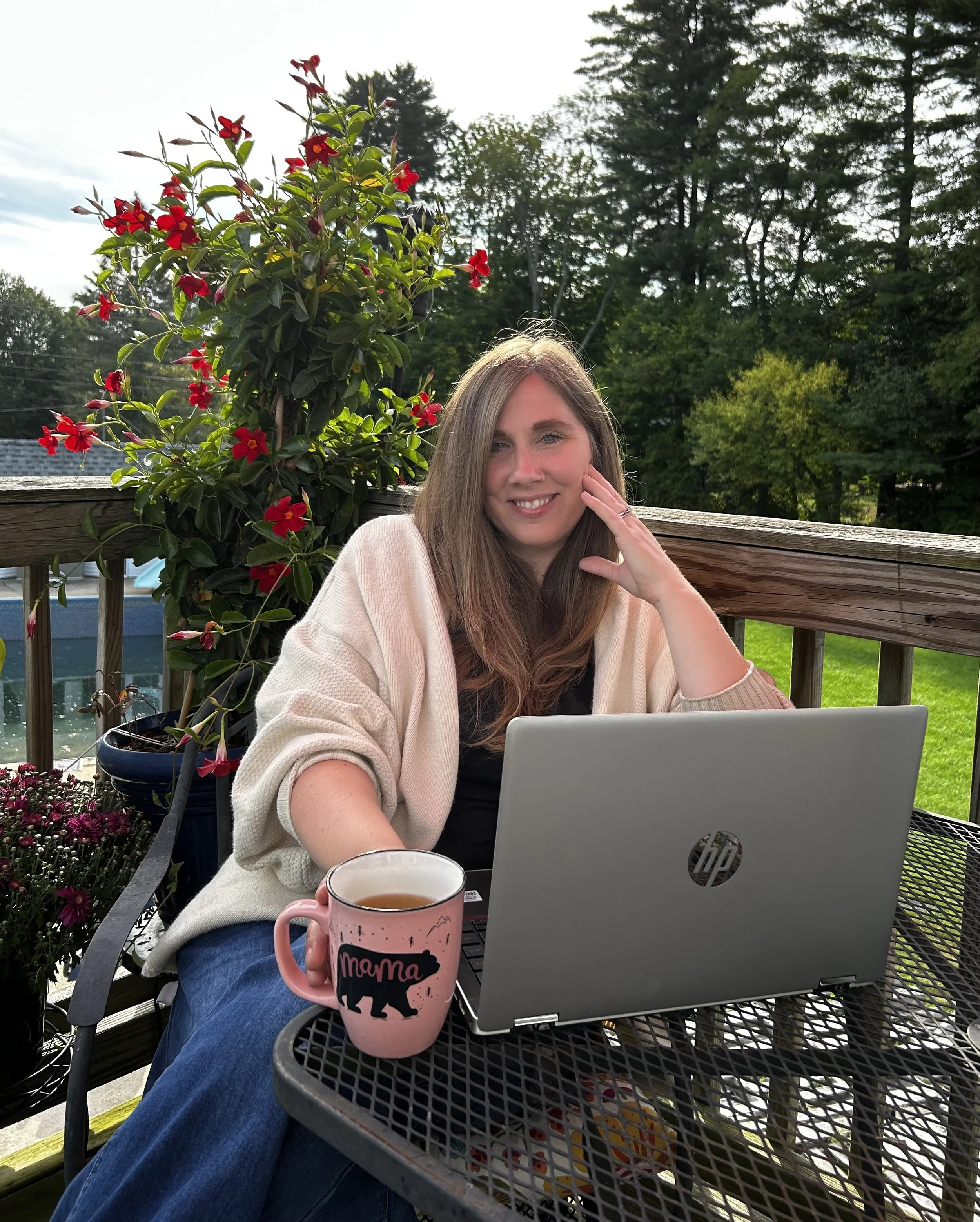 A woman sitting at a black metal table on a porch, smiling and resting her hand on her face. She has long, light brown hair and is wearing a light-colored cardigan over a black top. On the table in front of her is an open silver HP laptop and a pink mug with black and white text and a bear graphic. In the background, there is a wooden fence, a potted plant with red flowers, and a lush, green, wooded area with trees under a partly cloudy sky.
