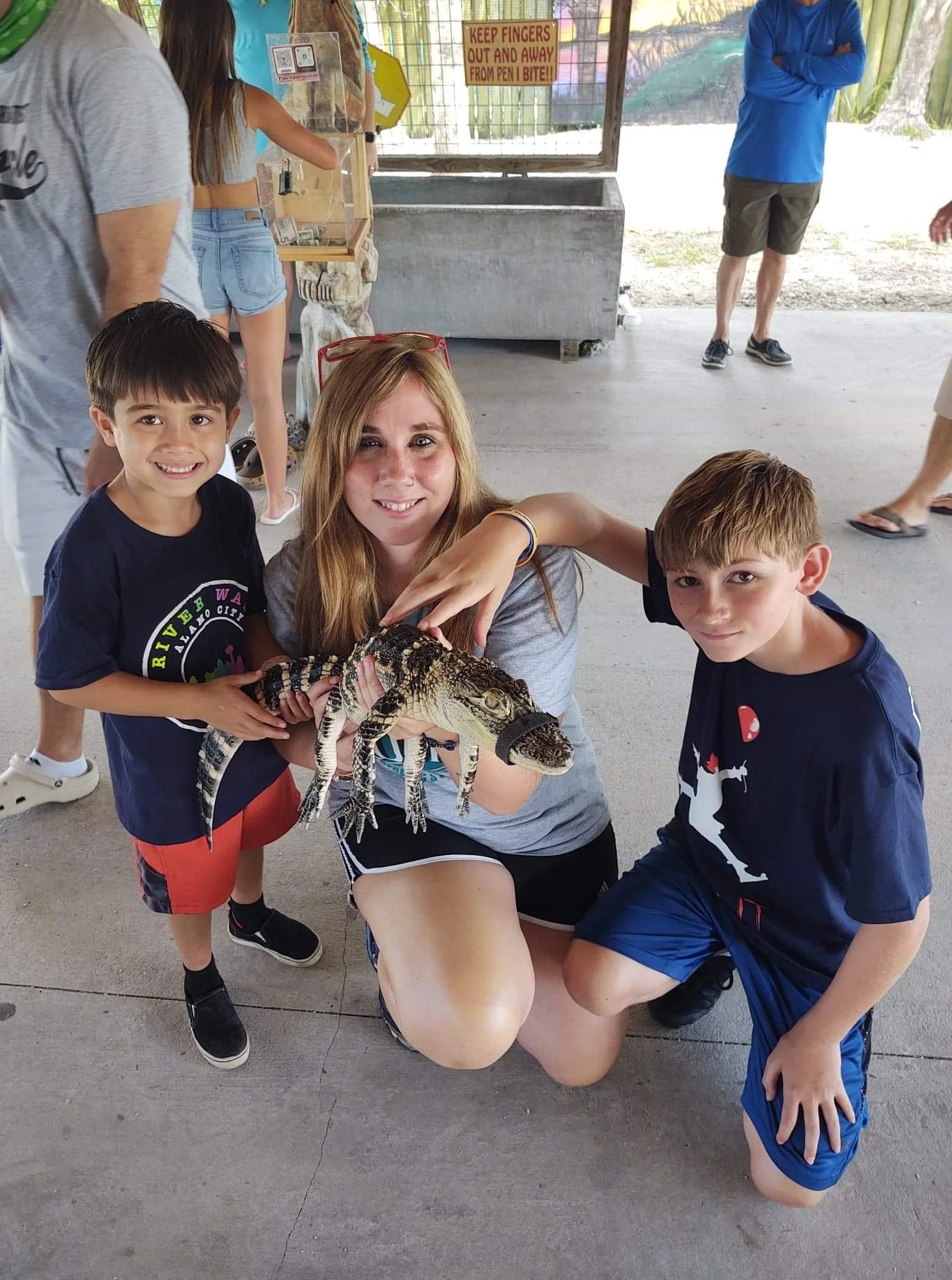 A group of children and a woman posing with a small alligator at an outdoor zoo or animal exhibit. The woman is kneeling, holding the alligator, while the kids stand around her. The background shows a fenced area with a sign that reads, 'KEEP FINGERS OUT AND AWAY FROM PEN! BITE!!'