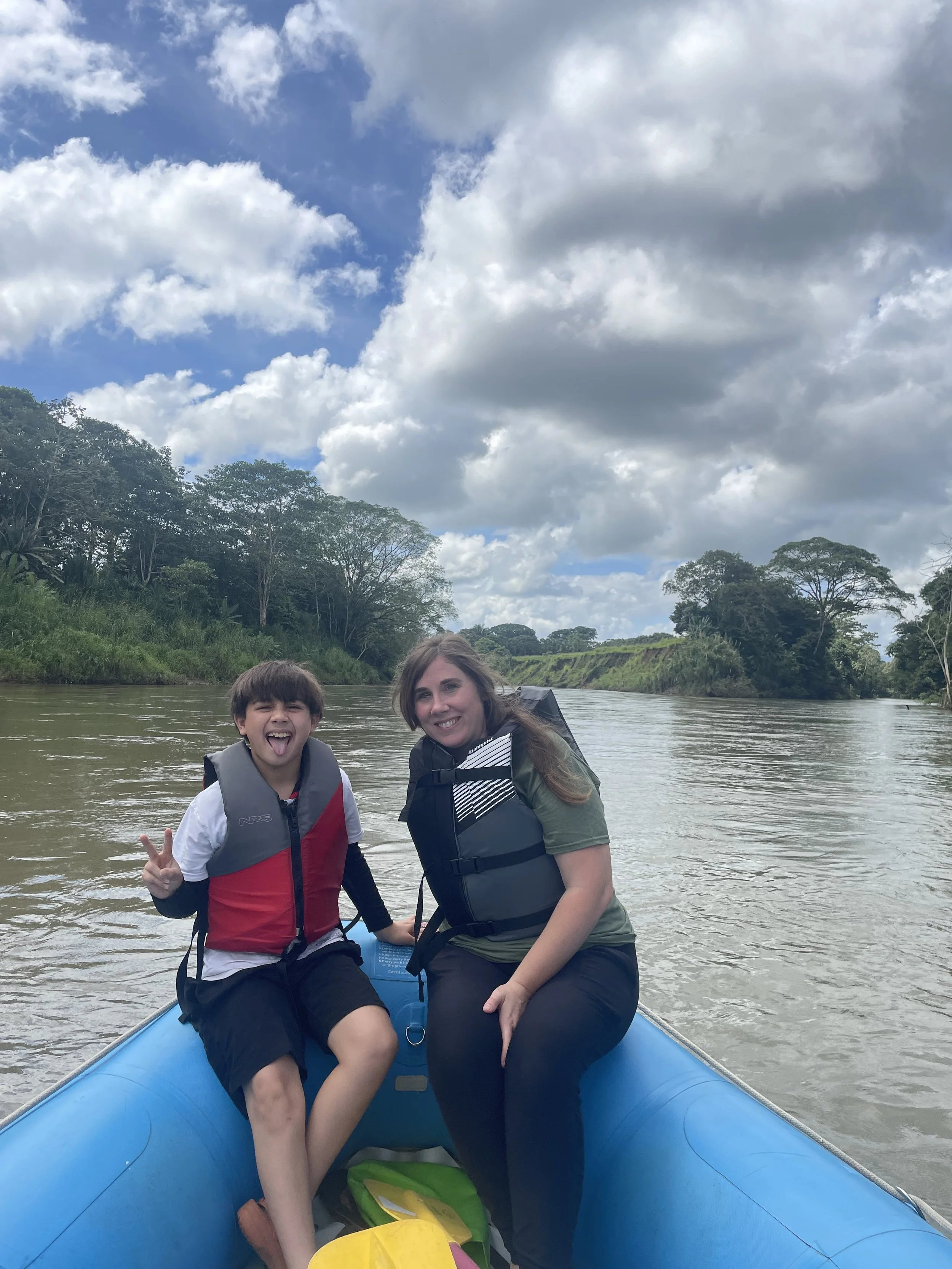 Two people, a boy and a woman, are sitting in a blue inflatable boat on a river surrounded by green trees and clouds in the sky. They are smiling, and the boy is making a peace sign with his hand.